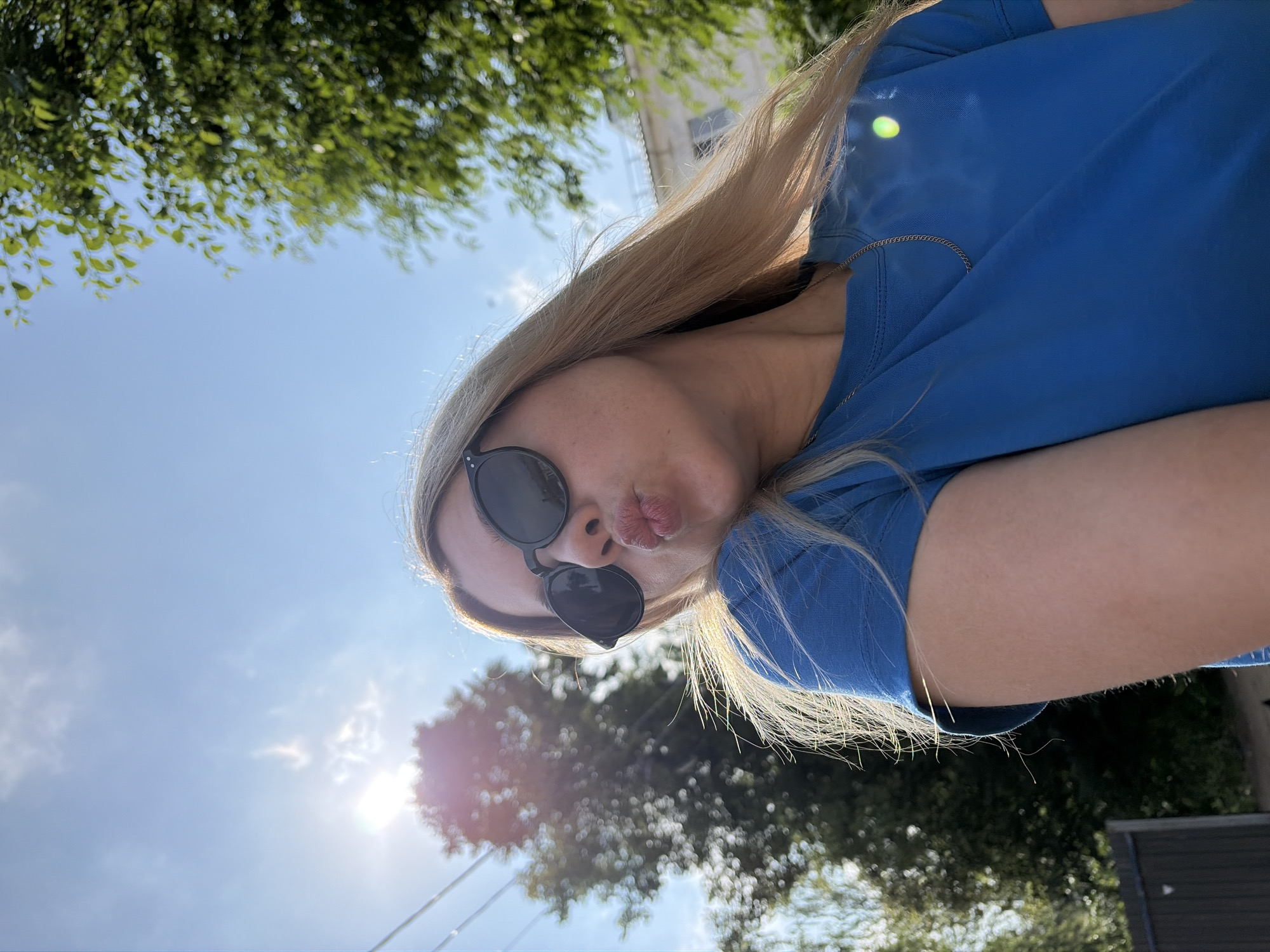 A woman with blonde hair and sunglasses is making a kissing face at the camera. She is wearing a blue t-shirt and a gold chain. The sun is shining brightly in a blue sky, with trees and a building visible in the background. The scene appears to be outdoors, possibly in a park or residential area. The location is identified as Chaikovsky, Russia, though no specific landmarks are discernible from the image itself to confirm this.