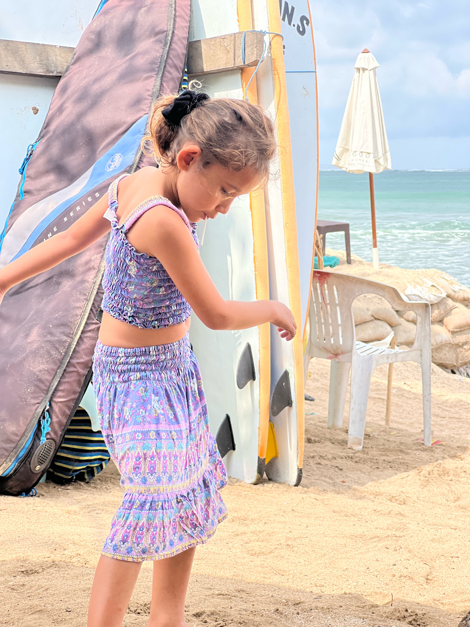This media file captures a young girl with blonde hair tied back with a black scrunchie, wearing a purple floral-patterned two-piece outfit consisting of a smocked crop top and a ruffled skirt. She is standing on a sandy beach, with her head bowed as if observing something on the ground.
Behind her, several surfboards are leaning against a structure. One surfboard is partially covered by a dark grey bag with blue trim and yellow accents. Another surfboard is white with orange edging and has the partial text "N.S" visible at the top. Further in the background, the ocean is visible with gentle waves under a bright, cloudy sky, suggesting a daytime scene. A folded white beach umbrella stands near the water, and two plastic chairs and sandbags are scattered on the sand.
The setting is clearly a beach in Kuta, Indonesia, indicated by the surfboards and the overall ambiance. The lighting suggests it is a sunny day. The girl's posture and focused expression hint at concentration or curiosity.