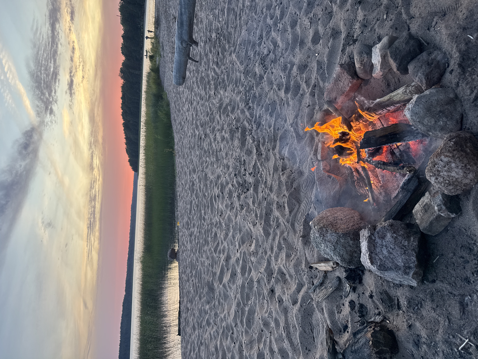 The image depicts a beach scene at sunset. In the foreground, a campfire burns within a ring of stones and bricks on the sandy shore. The fire consists of logs and branches, with flames and embers visible. Several rocks of varying sizes surround the fire pit. Beyond the sand, tall green reeds grow along the edge of a body of water, which reflects the sky. A dark, dense line of trees forms the horizon on the far side of the water. The sky displays a gradient of colors, transitioning from pink and orange near the horizon to lighter blues and whites higher up, with scattered clouds. The sandy ground shows footprints and the texture of the sand. In the distance, a large log lies on the beach. The overall scene conveys a tranquil, natural setting.