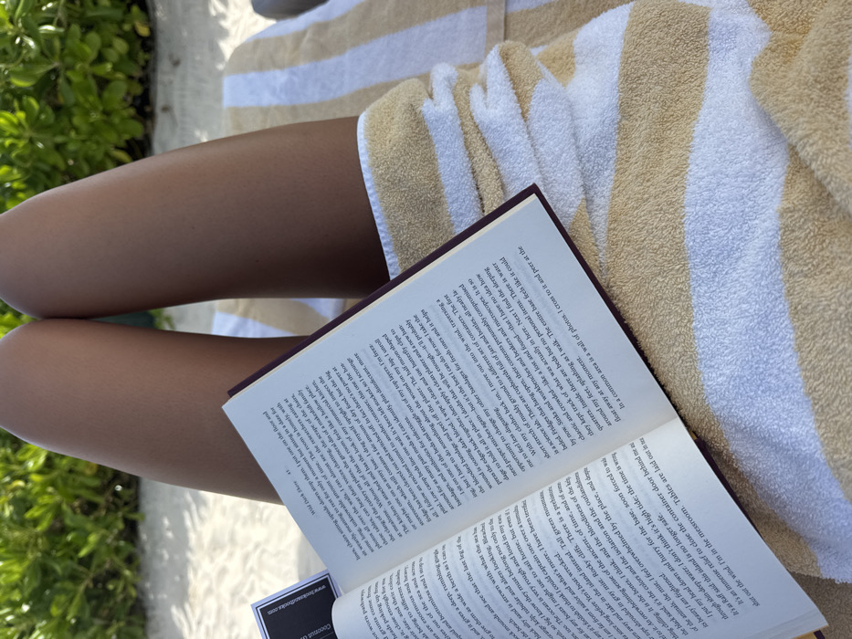 The image displays a close-up, downward-facing perspective of an individual reclining outdoors in Marina Maroma, Mexico. The lower torso and legs of a person with tanned skin are visible, extending upwards from the bottom of the frame. The individual is covered from the waist down by a horizontally striped beach towel in alternating white and light tan/gold bands. An open paperback book, featuring a dark maroon spine and white pages with black text, rests on the person's lap, its pages facing upwards. Page 41 is visible, displaying text and the partial title "Wild Dark Here." A small black rectangular object with white text, possibly "Coconut Club," is partially visible to the left of the book. The background consists of vibrant green foliage, indicative of a tropical or lush outdoor environment. The person is engaged in reading the book while reclined.