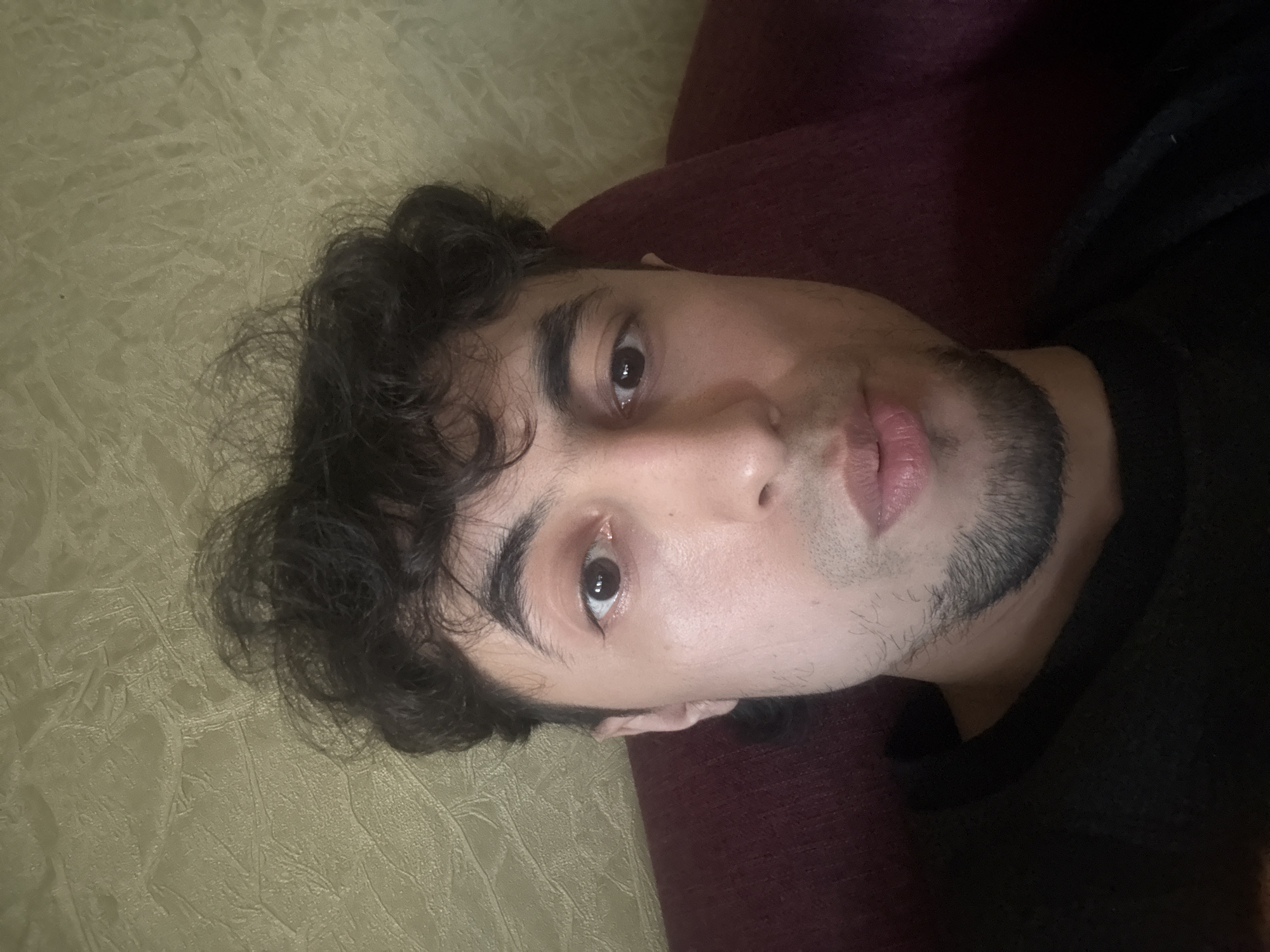 The media file is a close-up, high-angle shot of a young person lying down, with their head tilted to the left. The person has dark, curly hair and is wearing a dark-colored top, possibly a sweater. Their skin is fair, and they have dark eyebrows and a light beard. One eye is clearly visible, showing a dark iris. The person's lips are slightly parted, revealing a hint of pink. The background is divided into two distinct textures: to the left, there is a textured, light-colored surface, possibly a fabric or a wall covering with a subtle pattern. To the right, there is a darker, plush-looking material, likely upholstery from a couch or chair, in a deep red or maroon color. The lighting appears to be dim and artificial, suggesting the photo was taken indoors, possibly in the evening or at night. No text is visible in the image. The overall mood of the image is introspective or relaxed, given the subject's pose and the close framing.