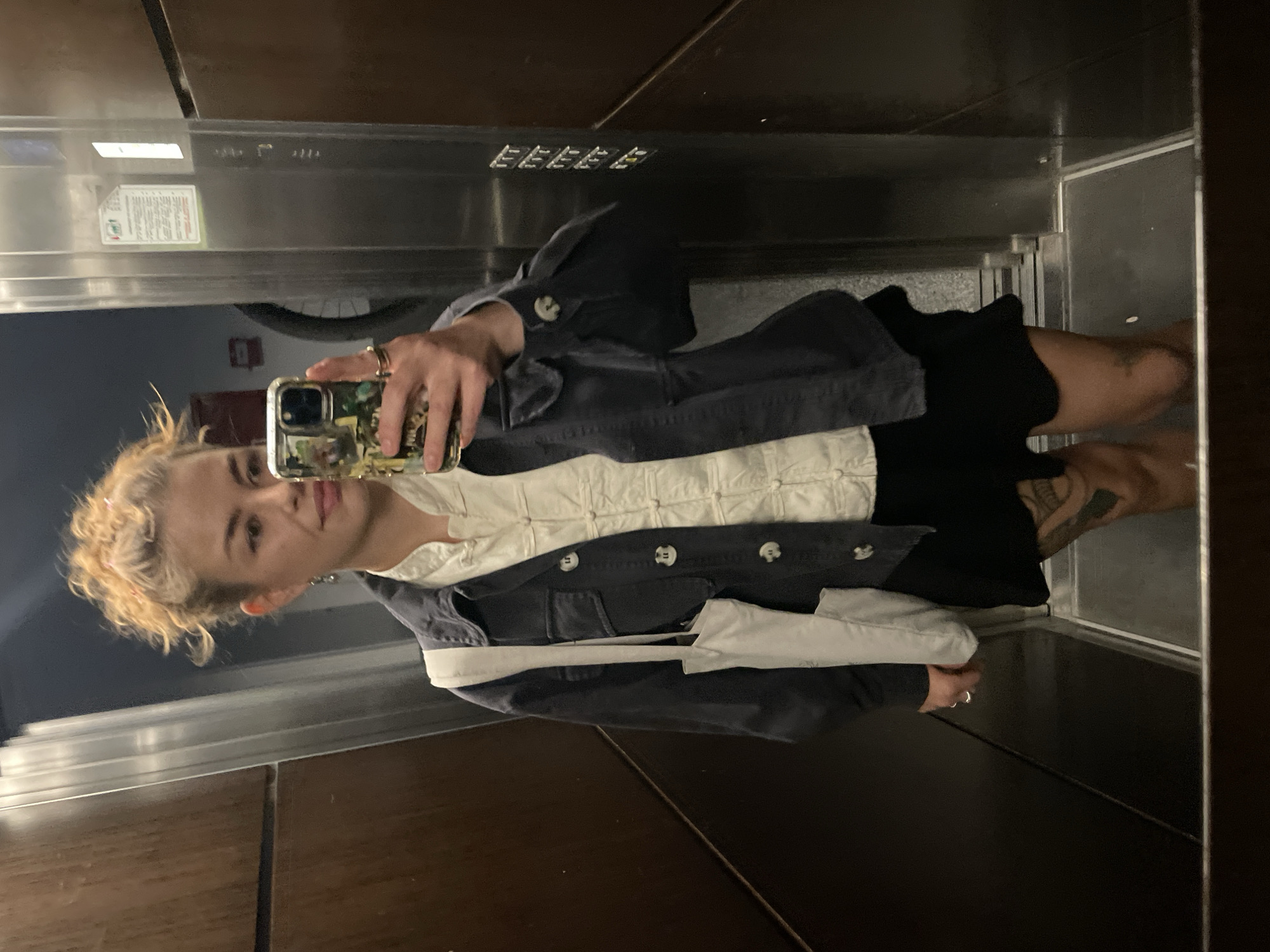 A young woman stands in an elevator, taking a mirror selfie with her smartphone. She is wearing a white, button-down top, a dark jacket, and a black skirt. She has blonde, curly hair pulled back in a messy bun. The elevator is enclosed with dark wood paneling and a stainless-steel ceiling with recessed lights and signage. The location is an elevator, likely in a building in Boreti, Montenegro.