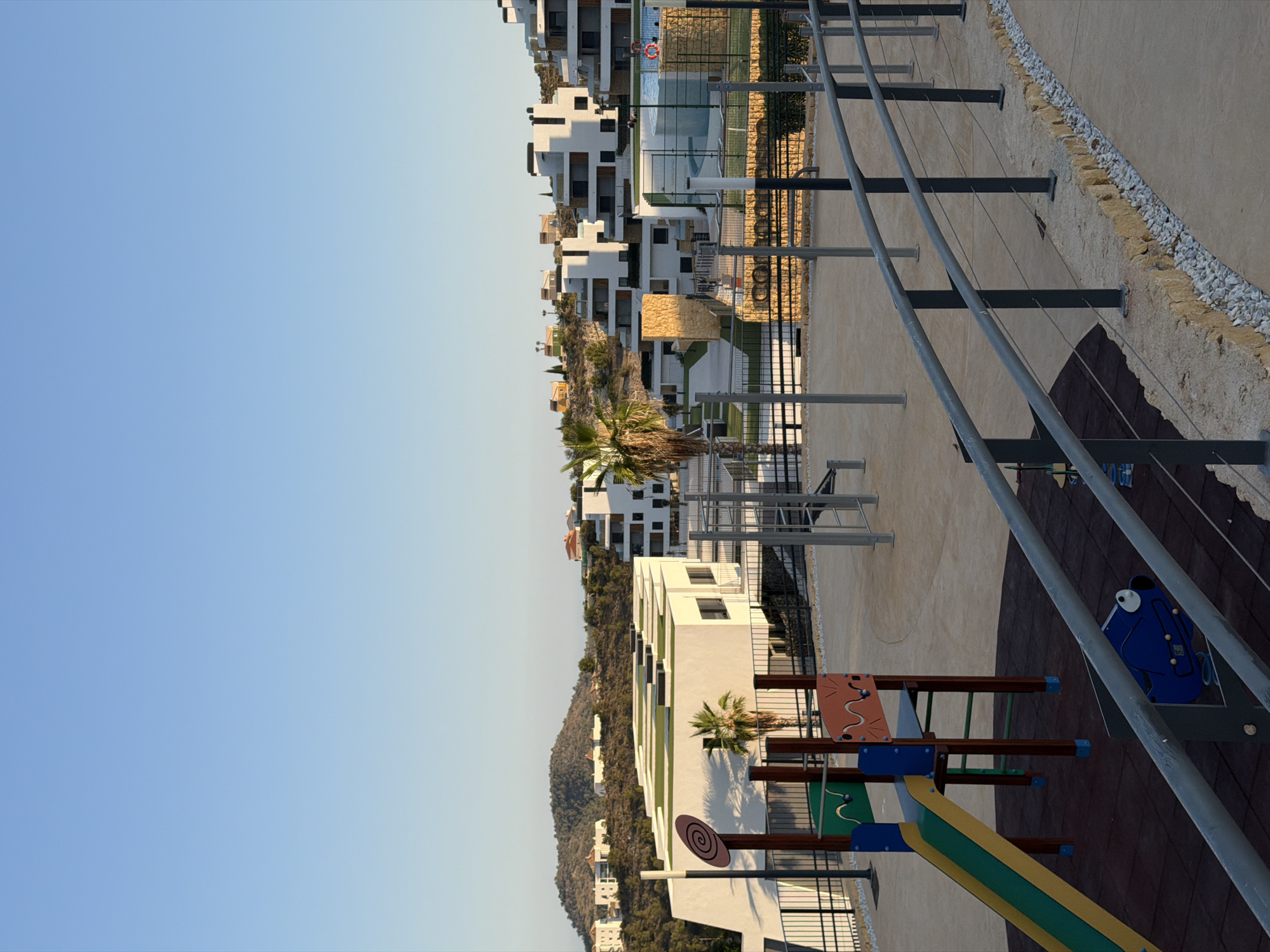 The image depicts an outdoor playground and sports facility located in the city of Finestrat, Spain. In the foreground, a colorful children's slide with green and yellow accents is visible, along with a blue ride-on toy shaped like a dog. A metal railing system curves along the right side of the frame, separating the playground from a walking path.
Behind the playground, a modern white building with a green rooftop is situated. Further in the background, a complex of white, multi-story buildings with geometric designs is built into a hillside. These buildings appear to be part of a residential or resort complex. To the left, a palm tree stands near the white building. A tennis court and swimming pool are also visible in the middle ground. The landscape is characterized by rolling hills covered with vegetation and scattered houses. The sky is a clear, pale blue, indicating daytime with fair weather. The overall scene suggests a well-maintained recreational area within a contemporary development.