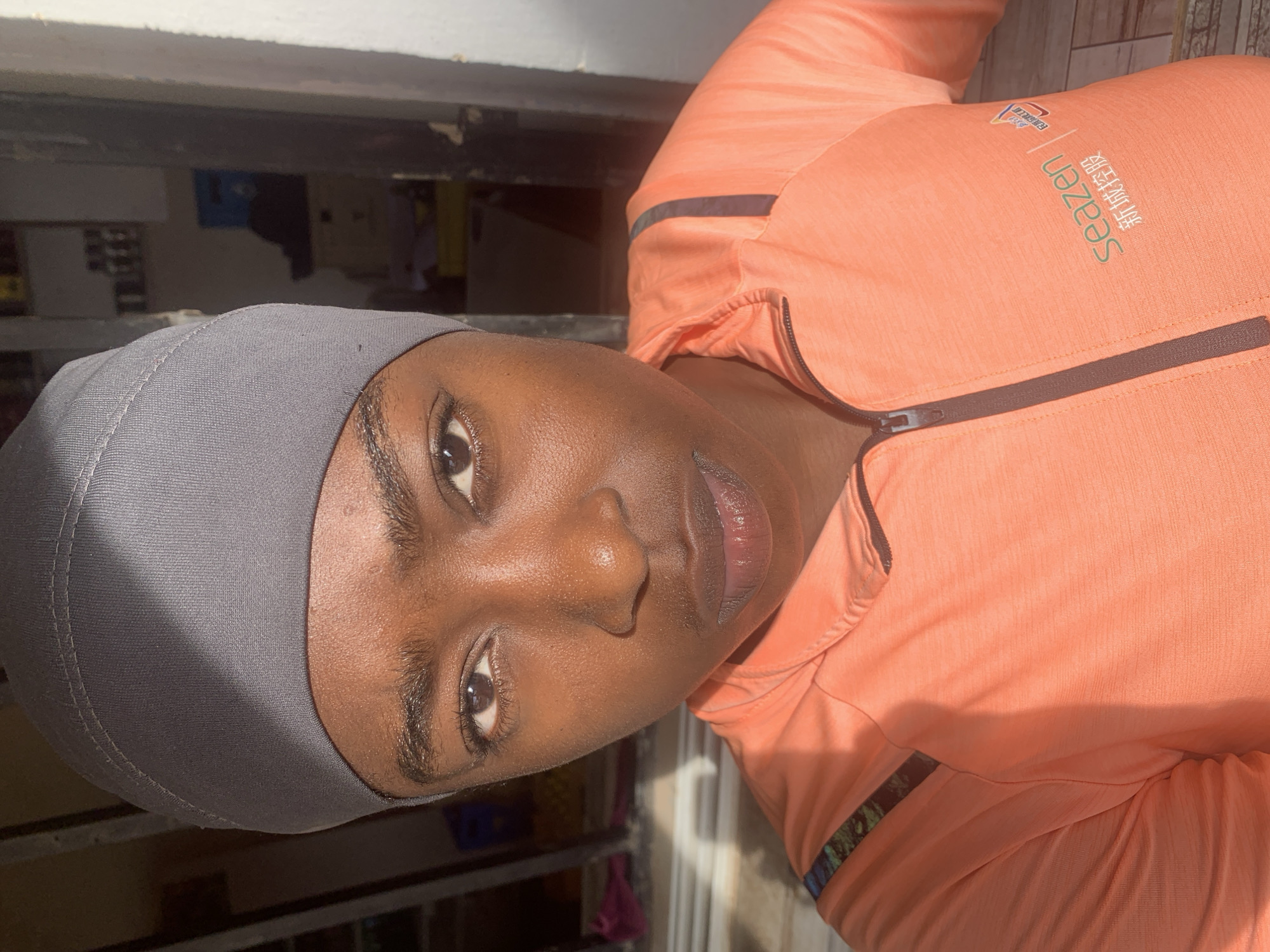 A medium-close shot depicts a dark-skinned individual with an apparent head covering, likely a hijab, with a light gray hue. The person is wearing an orange zippered jacket with black accents. The text "Seazen" and accompanying Chinese characters are visible on the jacket. The individual gazes directly at the camera with brown eyes. The background reveals a partially visible interior space with shelves and other objects, and possibly a doorway or opening. The image suggests an indoor setting, possibly in the city of New Stadium, Nigeria. No interactions or actions are evident other than the individual's gaze at the camera.
