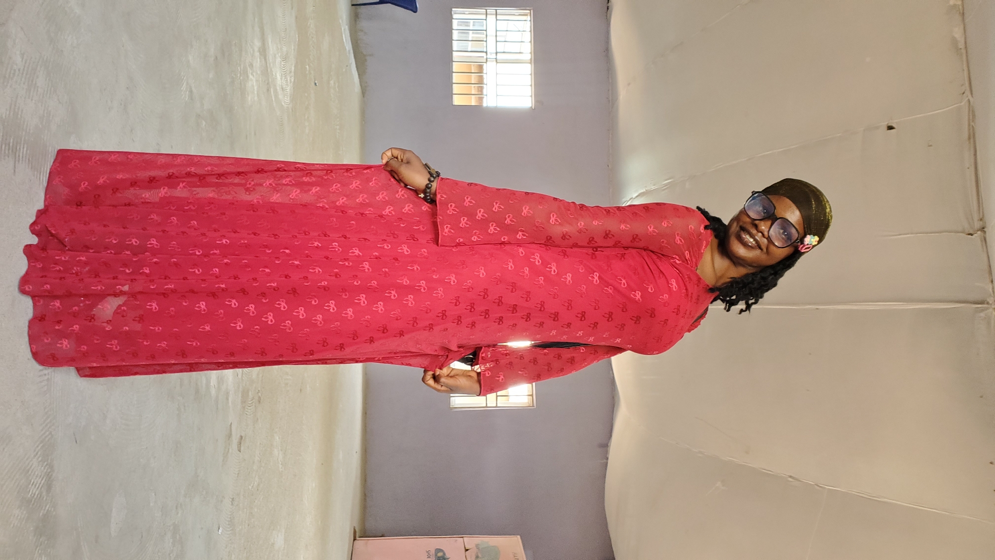 A woman is standing in an indoor space. She is wearing a long, red, flowy dress with a subtle bow pattern. She has on a dark-colored cap with some embellishments and large, black-rimmed glasses. Her hair is dark and braided, with some strands falling over her shoulders. She is smiling and looking towards the camera. Her right hand is holding the fabric of her dress. The environment appears to be a large, empty room with a light-colored floor and pale walls. There are windows with grates visible on the left wall, and a white, tent-like ceiling. A pink cabinet with some writing is visible on the far right. The image appears to have been taken in Jalingo, Nigeria, likely in a community hall or event space.