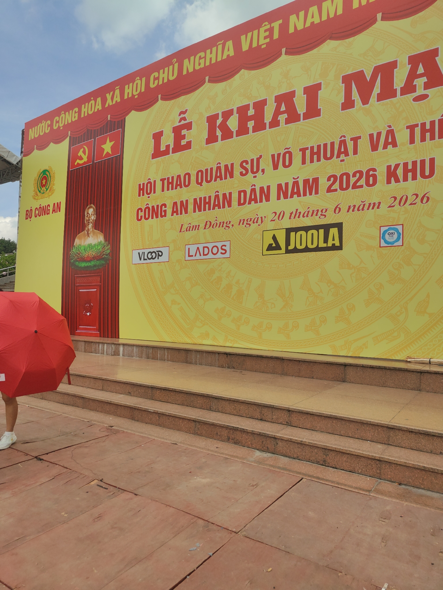 This image captures a portion of a large yellow banner displayed outdoors under a bright sky with scattered clouds. The banner features prominent red text announcing an event. Dominating the upper right portion of the banner is the bold red text "LỄ KHAI MA" which appears to be the beginning of the title of the event. Below this, smaller red text reads "HỘI THAO QUÂN SỰ, VÕ THUẬT VÀ THI" followed by "CÔNG AN NHÂN DÂN NĂM 2026 KHU". Further down, in smaller text, it says "Lâm Đồng, ngày 20 tháng 6 năm 2026".
To the left of the banner, a portion of a building or structure is visible, with a red curtain and a portrait of Ho Chi Minh. The words "BỘ CÔNG AN" (Ministry of Public Security) are printed in red on a green circular emblem to the left of the portrait. Two flags are displayed above the portrait: a red flag with a yellow hammer and sickle, and a yellow star on a red field.
In the foreground, a person is partially visible on the left, holding a red umbrella. The ground in front of the banner consists of weathered reddish-brown wooden planks. A series of tiered steps, appearing to be made of granite or a similar stone, lead up to the area where the banner is displayed. The overall scene suggests an outdoor public event, possibly in Da Lat, Vietnam, given the mention of "Lâm Đồng" which is a province containing Da Lat.
