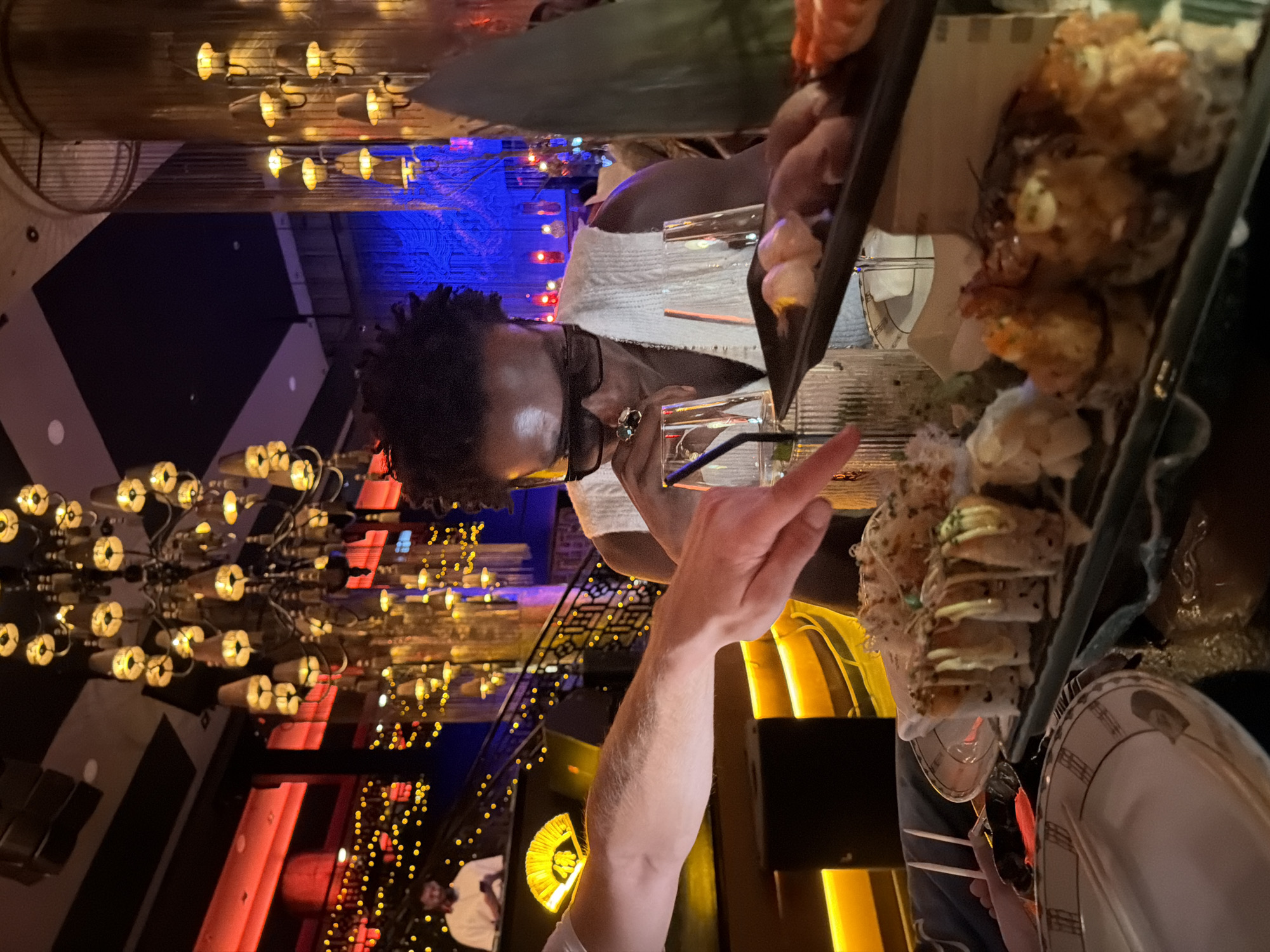 The image depicts a person seated at a table in a dimly lit, upscale restaurant. The person is wearing a white sleeveless top and large sunglasses. They are looking down at a plate of sushi in front of them, with their right hand near their mouth and a ring on their index finger. Another person's arm and hand are visible in the foreground, with the hand pointing towards the sushi. The table is laden with various dishes of sushi and drinks. The restaurant's interior is elaborate, with a large, ornate chandelier hanging from the ceiling and decorative lighting creating a vibrant, atmospheric ambiance. The background shows a staircase with red and yellow lights, and blue lighting further back. The overall setting suggests a luxurious dining experience. There is no information in the image to indicate that the location is Marrakesh, Morocco.