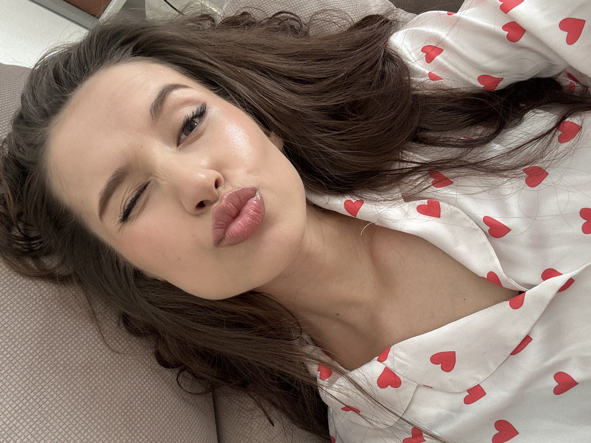 A young woman with long, dark brown hair is lying down, possibly on a couch or bed. She is wearing a white satin pajama top with red hearts. She is winking one eye and pursing her lips in a kiss. The background is blurred, but it appears to be an interior setting. The location is not discernible from the image.