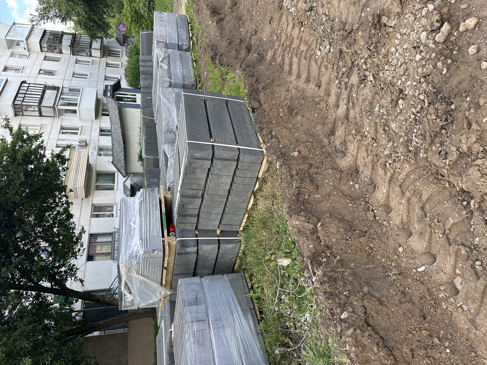 The image captures an outdoor urban scene, likely depicting a construction or renovation site adjacent to a residential building. The perspective is slightly tilted.
In the foreground and midground on the right, a strip of disturbed, brown earth mixed with gravel is visible, showing clear tire tracks, suggesting a prepared bed for a pathway or trench. Adjacent to this, a patch of green grass runs parallel to the dirt.
Stacked neatly on wooden pallets on the grass, in the midground, are numerous dark gray paving stones or tiles. Most stacks are wrapped in clear plastic sheeting and secured with white straps, indicating they are new construction materials awaiting installation.
On the far left, a multi-story residential building dominates the background. Its facade is light-colored, featuring multiple windows and various types of balconies; some are enclosed with glass, while others have dark metal railings. An air conditioning unit is visible on the building's exterior. A large, leafy green tree partially obstructs the view of the building on the left side.
At the ground level of the building, an entrance with a small canopy is visible. Text on the canopy reads "ДОБРО ПОЖАЛОВАТЬ" (Dobro Pozhalovat', meaning "Welcome"). Below this, on the entrance door, the text "24 ЧАСА" (24 Chasa, meaning "24 Hours") is discernible.
The overall scene suggests ongoing or imminent landscaping or construction work, likely for a new sidewalk or pedestrian area using the paving stones. The weather appears to be clear or partly cloudy daytime, with bright, even illumination. The building's architecture is characteristic of residential blocks found in cities like Chaikovsky, Russia. No people are visible.