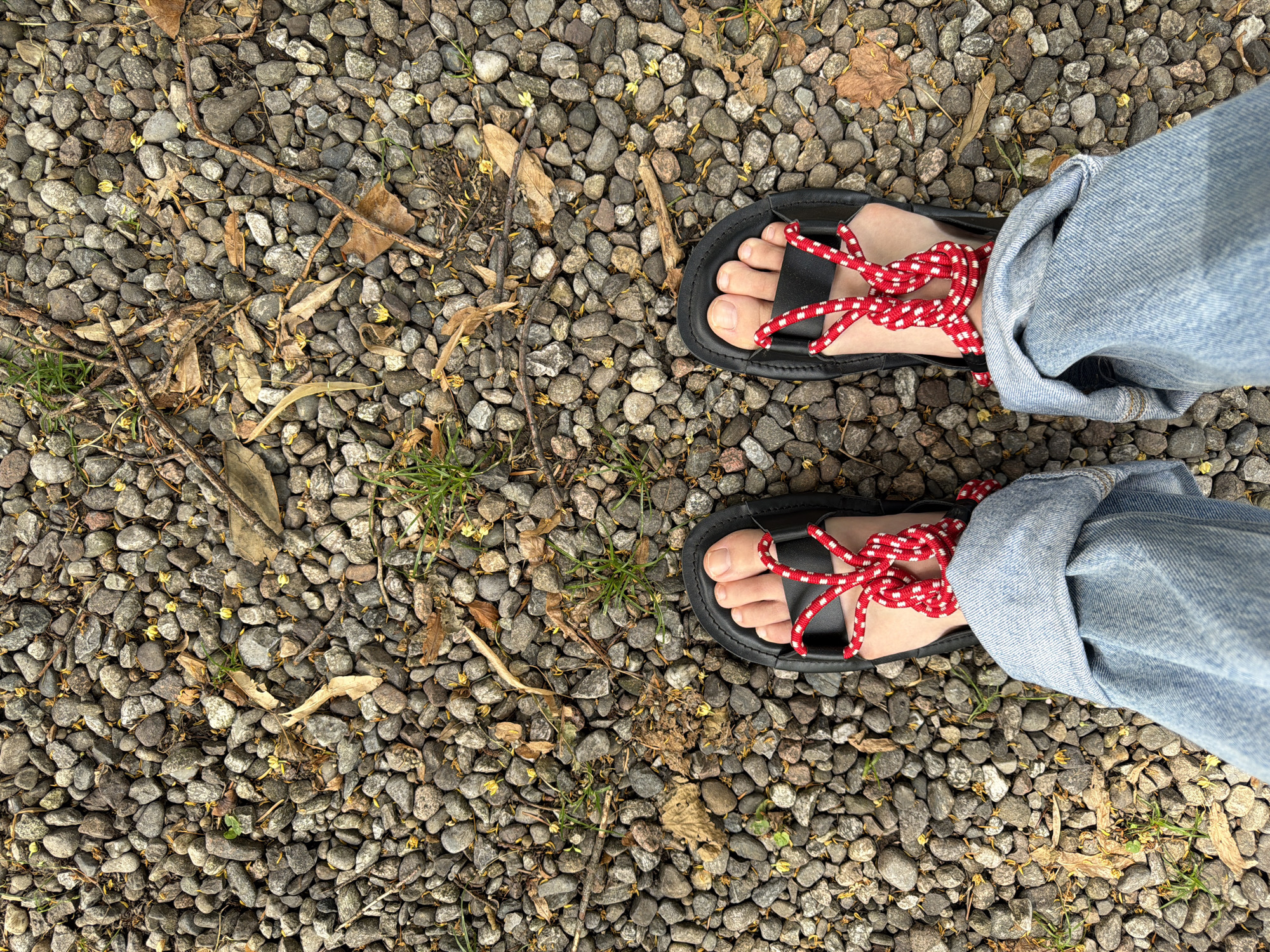 A first-person perspective shot shows a pair of feet wearing black sandals with red and white patterned laces, standing on a gravel-covered ground. The gravel consists of small, grey, and reddish-brown stones, interspersed with dried leaves, twigs, and a few blades of green grass. The individual is wearing light blue denim jeans, the cuffs of which are rolled up and resting over the sandals. The scene appears to be outdoors, possibly in Penzano, Italy, with the gravel pathway indicating a potentially natural or semi-natural setting.