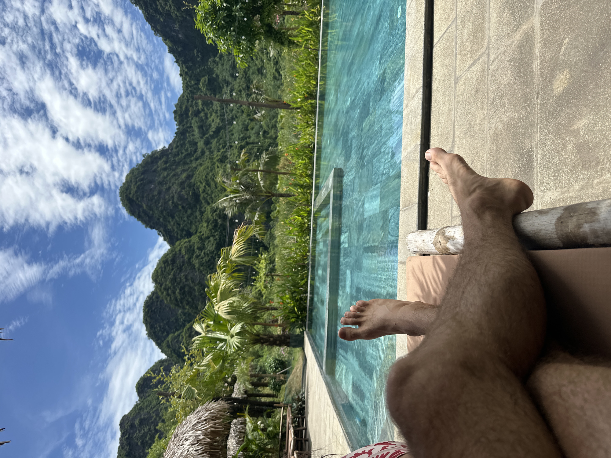 The image depicts a person's legs and feet resting on a lounge chair beside a swimming pool. The pool water is a clear turquoise color, with visible tile patterns on the bottom. In the background, lush green mountains rise steeply, covered in dense vegetation. The sky is a vibrant blue with scattered white, wispy clouds. Palm trees and tropical plants surround the pool area, and a thatched-roof structure is visible to the left. The scene suggests a relaxed vacation setting in a tropical or subtropical location. The location is identified as Nam Hoa Lư Ward, Vietnam.