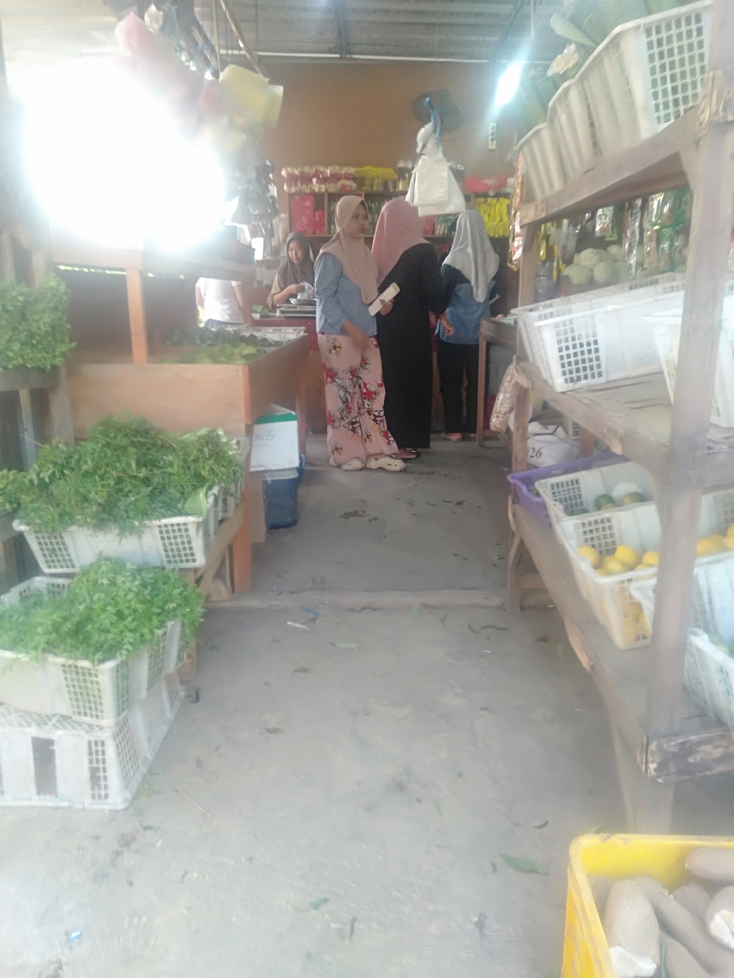 The image depicts an outdoor market scene in Sukadami, Indonesia, with a focus on fresh produce. In the foreground, several white plastic crates are filled with green leafy vegetables, some stacked on top of others. Behind these crates, a wooden stand displays more vegetables in similar crates. To the right, a wooden shelving unit holds more crates containing fruits, including lemons and possibly oranges. Several women are present in the market. One woman in a light pink hijab and a blue shirt, wearing floral patterned pants, stands near the produce. Another woman in a dark hijab and black clothing is also visible, as is a third woman in a grey hijab and a blue top. The ground is paved and appears somewhat weathered. The overall impression is of a busy, open-air market with a variety of fresh goods for sale.