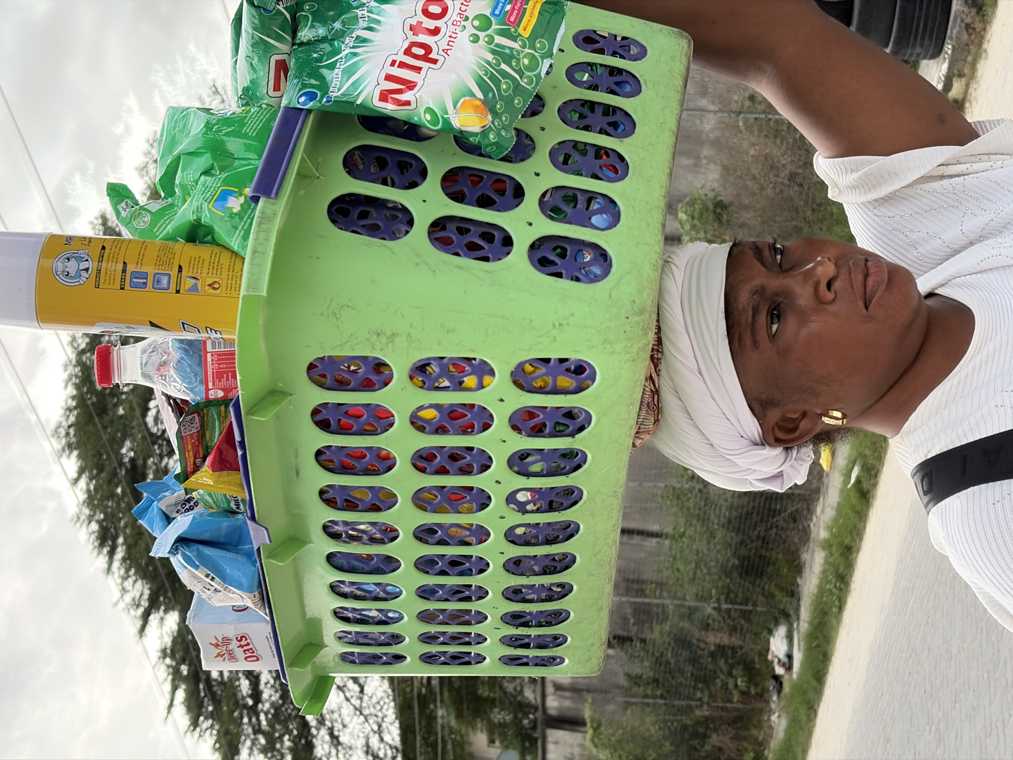 A woman in Igboefon, Nigeria, balances a large green plastic basket filled with various goods on her head. The basket contains items such as "Cheer-up Oats," blue and yellow packaged snacks, and multiple green packets of "Nipton Anti-Bacterial" detergent. A yellow spray can and a clear bottle of liquid are also visible amongst the items. The woman, wearing a white headscarf and a white patterned top, has a serious expression as she walks outdoors. The background shows a paved road, a chain-link fence, and some vegetation, suggesting an urban or semi-urban environment. The sky is overcast, and the lighting indicates it is daytime.