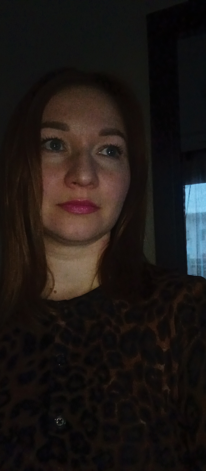 A woman with red hair, fair skin, and blue eyes is visible in the image. She is wearing a dark, animal print top with buttons. Her lips are coated in pink lipstick. The environment is dimly lit, suggesting an indoor setting, possibly near a window, as there's a hint of light from outside. The location appears to be a city with the name "Unknown" listed twice, likely due to a lack of specific information.