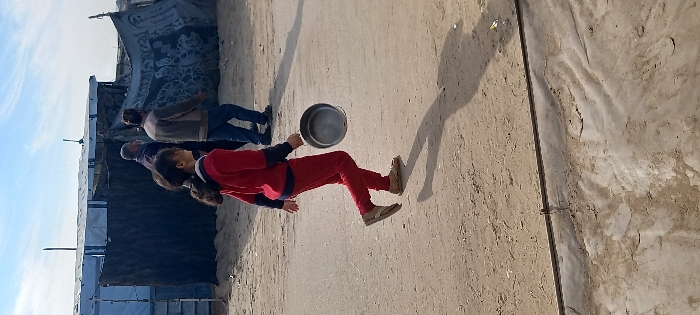 The scene depicts an unpaved, sandy-dirt pathway in Khan Yunis, Palestinian Territory, under a clear blue sky with some scattered clouds. Three individuals are walking from left to right across the frame. In the foreground, a young person, possibly a girl, with dark hair in a ponytail, walks wearing a red long-sleeved shirt with navy accents, red pants, and tan flip-flop sandals. She carries an empty silver-colored metal pot by its handle in her right hand, which swings slightly. Her long shadow extends diagonally forward on the ground. In the midground, two adult males follow, walking in the same direction. The man on the left wears dark clothing and has a beard. The man on the right wears a brown/grey top and blue jeans. In the background, makeshift shelters or tents are visible, constructed from light blue and dark tarpaulins, supported by poles. A dark, thin line, possibly a pipe or cable, runs horizontally across the lower portion of the sandy ground.