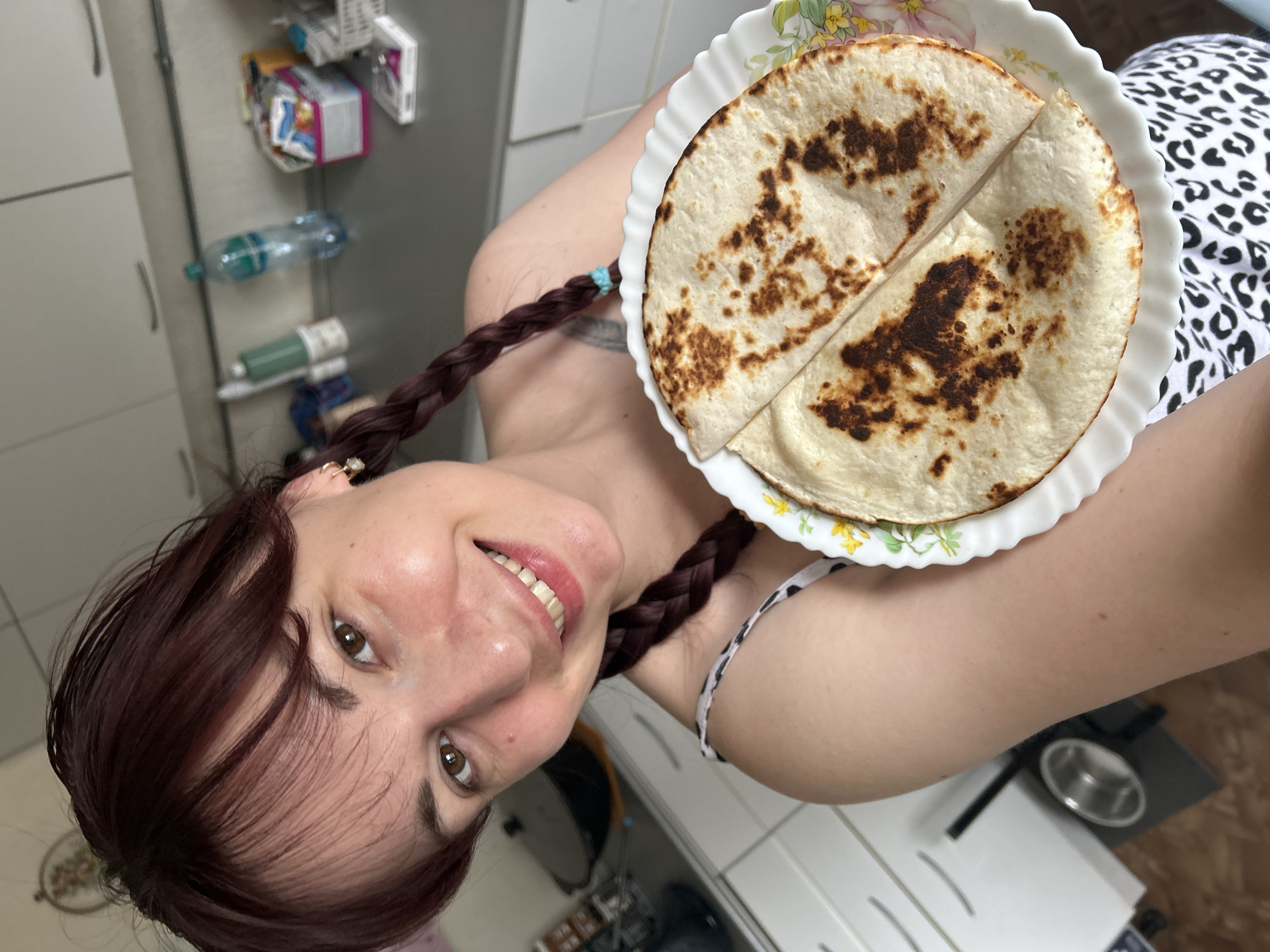 A young woman with dark red hair styled in two braids is holding a white plate with two folded flatbreads on it. She is smiling and looking at the camera. She is wearing a white top with a black and white leopard print. The background appears to be a kitchen with white cabinets and a countertop. A bottle of water and some containers are visible on the countertop. The floor is tiled in shades of brown and beige. A stainless steel bowl is on the floor in the lower left of the image. The scene is brightly lit, suggesting it is daytime. The location is identified as the city of Фоки, Russia.