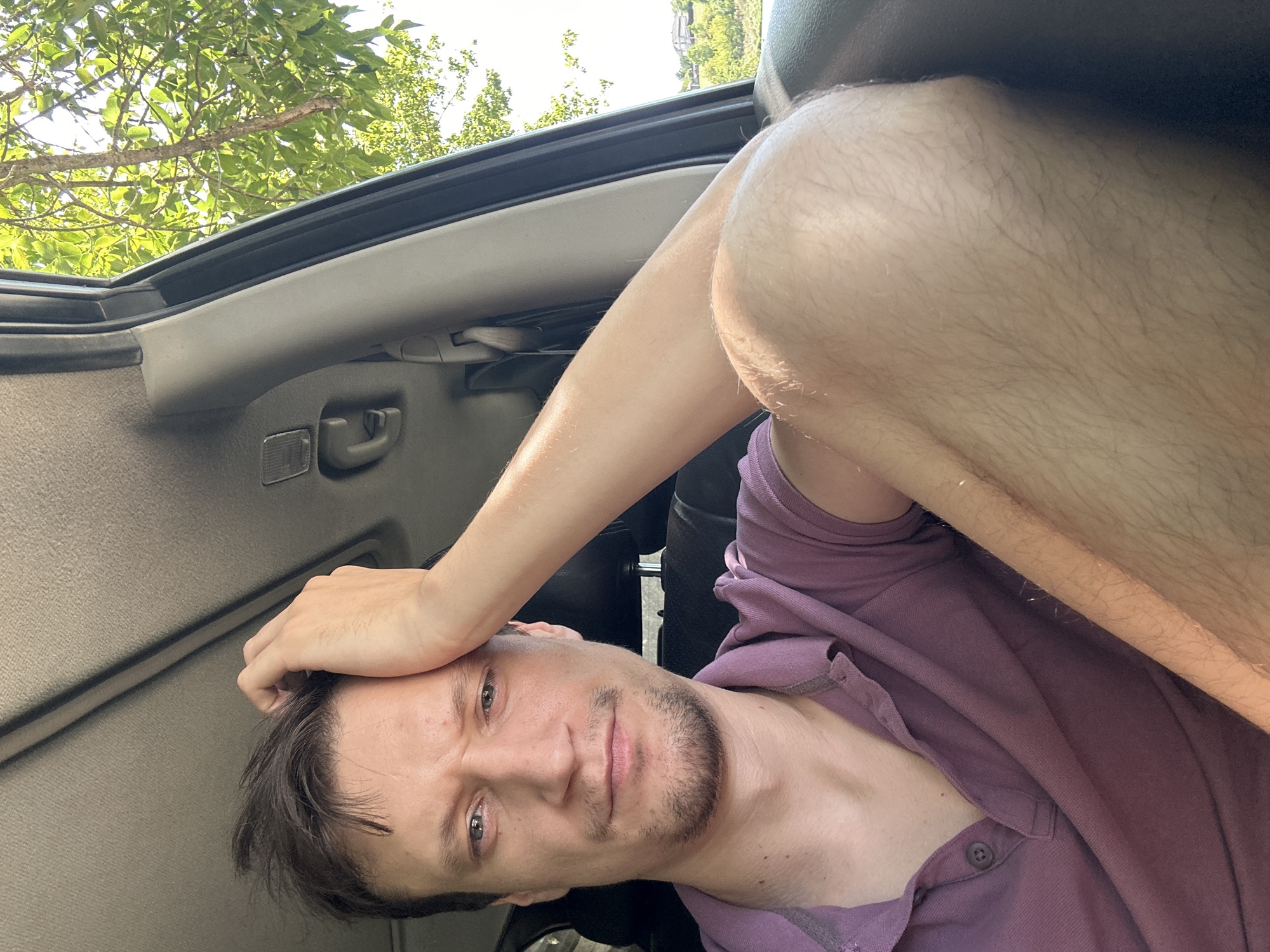 A man is seated inside a car, his left hand resting on his forehead. He is looking directly at the camera with a neutral expression. He is wearing a purple polo shirt. The interior of the car is visible, including the ceiling and a portion of the passenger seat. Outside the car window, trees and a clear sky are visible. The scene appears to be a sunny day. The location is specified as Makiv, Ukraine.
