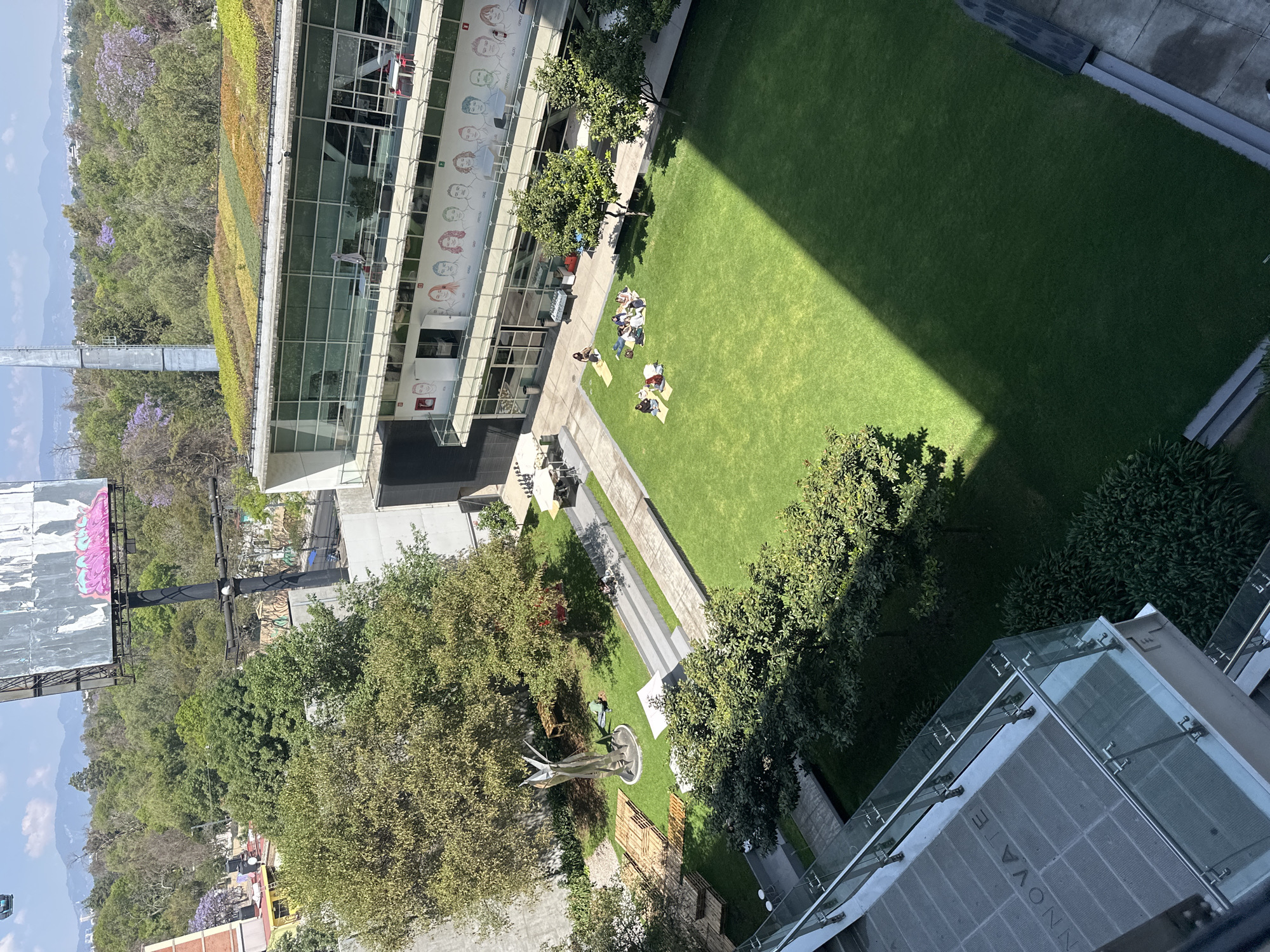The image captures an elevated view of a lush, green urban landscape within Mexico City, Mexico. A building with a green roof and a wall adorned with caricatures flanks the upper left. Adjacent to the building is a manicured lawn, where a small group of people are gathered, some sitting and others lying down. Shadows cast across the lawn indicate the time of day and position of the sun. Further down and to the left, a statue stands on a smaller patch of grass surrounded by trees. A large billboard is visible in the distance, along with the city skyline. The perspective suggests the image was taken from a high vantage point within a nearby structure.