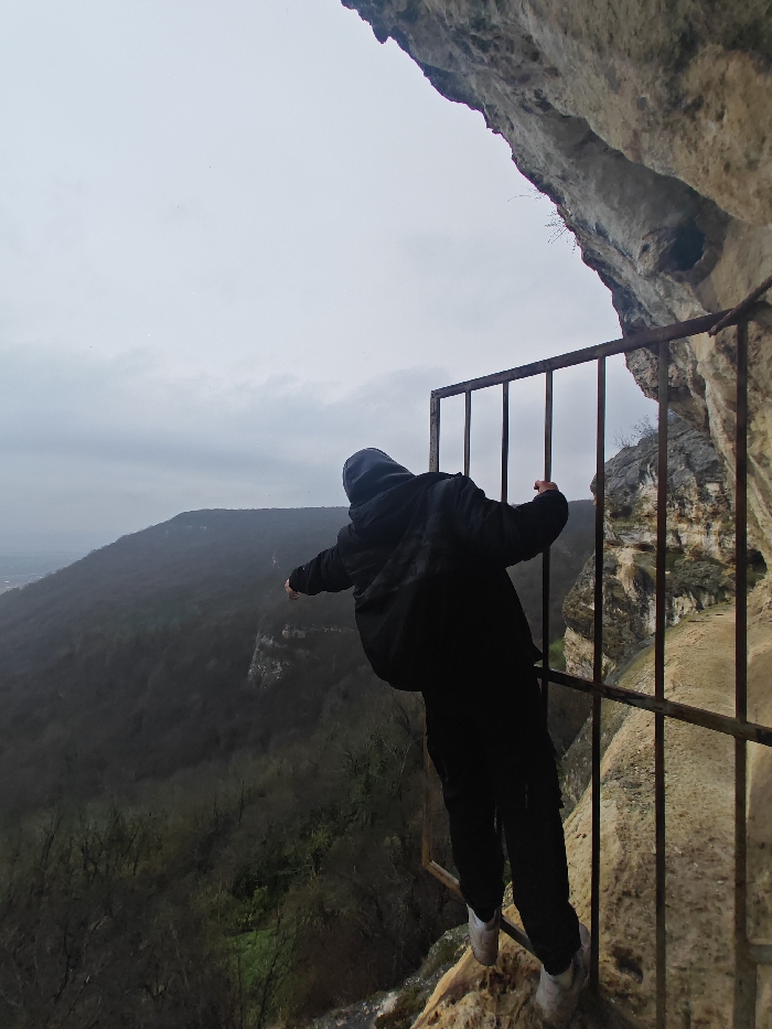 A single individual is observed on a narrow, uneven rocky ledge on a high-altitude rock formation on the Shumen Plateau, Shumen, Bulgaria. Positioned with their back to the viewer, the person is dressed in a dark hooded jacket, dark trousers, and light-colored athletic shoes. The individual is leaning outward over a precipice, gripping a rusty metal barrier composed of multiple vertical bars and horizontal cross-members, which is secured against the rock face on the right side of the frame. The rocky overhang above and to the right shows natural erosion. Below the ledge, a steep drop-off descends into a heavily forested valley, where dense, dark-green to brown deciduous trees are visible. An overcast, light-grey sky dominates the upper portion of the image, with a distant urban settlement faintly visible on the left horizon.