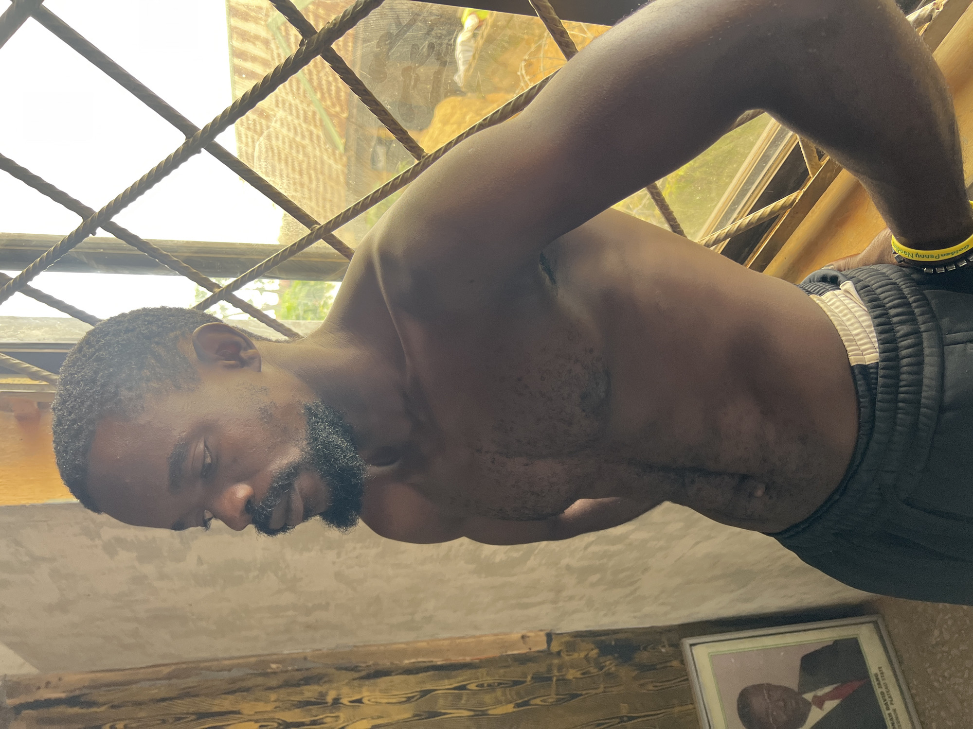 A shirtless Black male with dark skin, short black hair, and a goatee stands in front of a window with metal bars. He is wearing dark pants with a white stripe. To his left, a framed portrait of a Black man in a suit and red tie is visible. The background outside the window appears to be a residential area. The overall lighting suggests it is daytime. The image was taken in Jos, Nigeria.