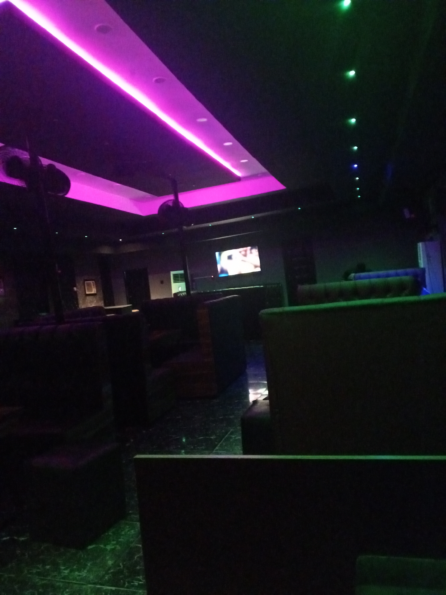 The image depicts the interior of a dimly lit space, possibly a nightclub or lounge, in an unknown location. The lighting is primarily provided by a strip of bright pink lights on the ceiling, along with a line of green and blue lights. Dark upholstered seating is visible throughout the room, with a television screen in the background displaying an image. The floor has a reflective quality, with a marble-like pattern. Two dark objects resembling speakers or fans are visible suspended from the ceiling.