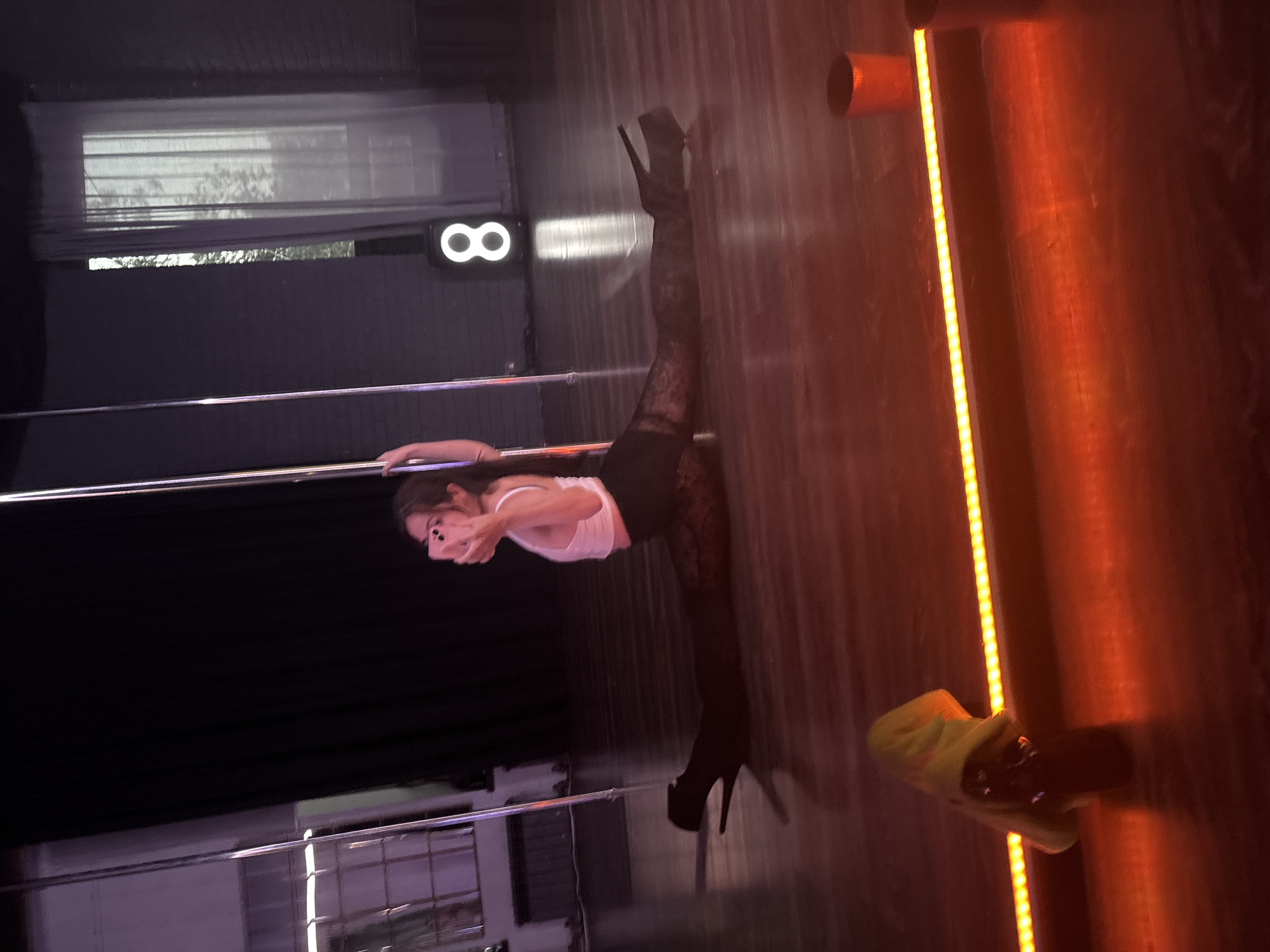 A person is performing a split on the floor in a dance studio. The person is wearing a white crop top, black patterned tights, and black high-heeled boots. They are holding a pink smartphone and taking a selfie. There are two poles on either side of the person, suggesting a pole dancing studio. In the background, there is a window with sheer curtains and a sign that reads "8". The floor appears to be made of wood. There is a yellow towel-like object on the floor in the foreground, and an orange light fixture runs along the bottom of the frame. The location is identified as a dance studio in Naberezhnye Chelny, Russia.