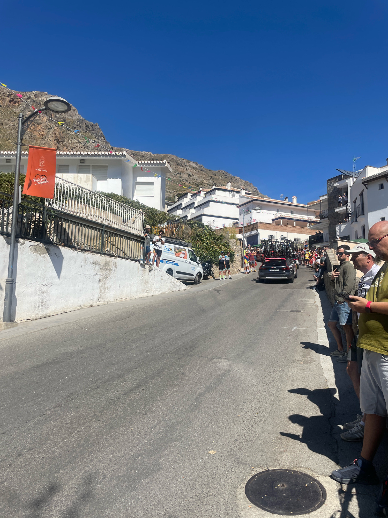 In Güéjar Sierra, Spain, on a sunny day, a street lined with white buildings and a mountain backdrop is filled with spectators awaiting an event. A black car with a rack of bicycles on top drives down the street, followed by a white van and a crowd of people. Spectators stand along the side of the street, some holding phones, to watch the procession. Decorative flags are strung across the street, and a lamppost with an orange banner hangs on the left side of the road.