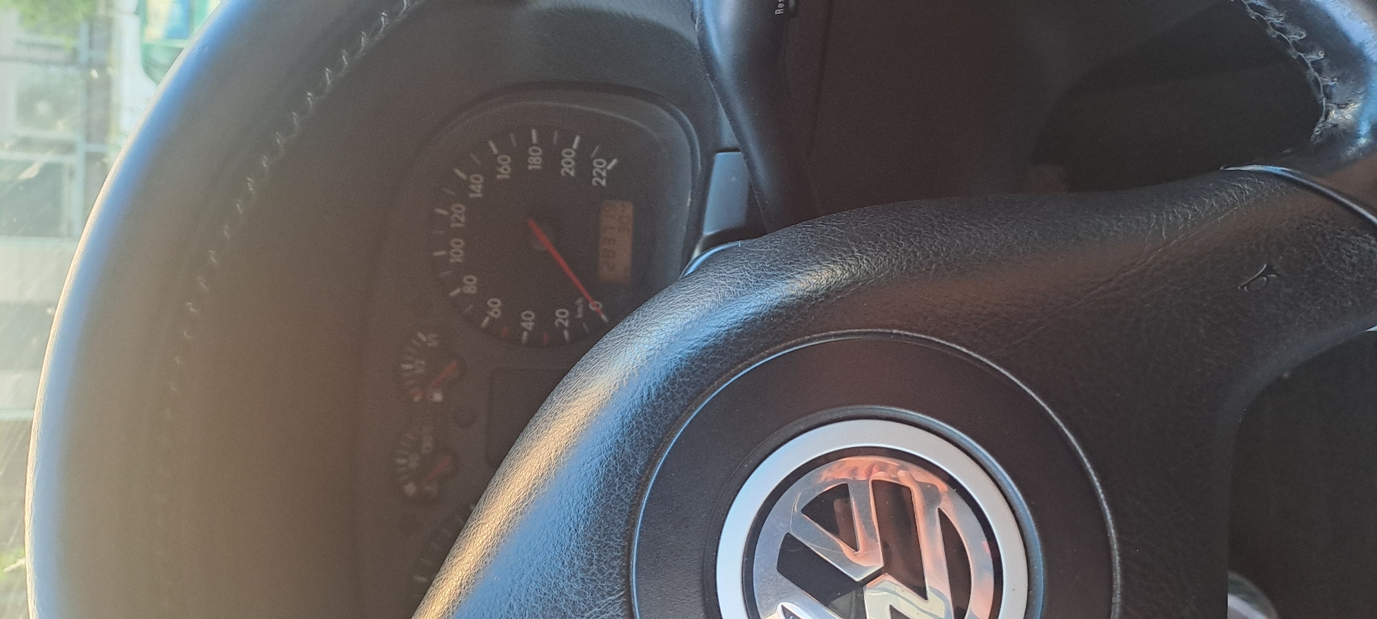 The image shows a close-up view of a car's steering wheel and dashboard. The steering wheel is black and textured, with a silver Volkswagen logo in the center. The dashboard features a speedometer and other gauges. The speedometer shows a speed of approximately 20 km/h, and the odometer reads 283119 km. The image appears to have been taken inside a car parked on a street in Burgas, Bulgaria, as indicated by the reflection of buildings and greenery in the steering wheel, which are consistent with an urban environment in Burgas. The lighting suggests it is daytime, possibly late afternoon due to the warm sunlight. There are no people visible in the image.