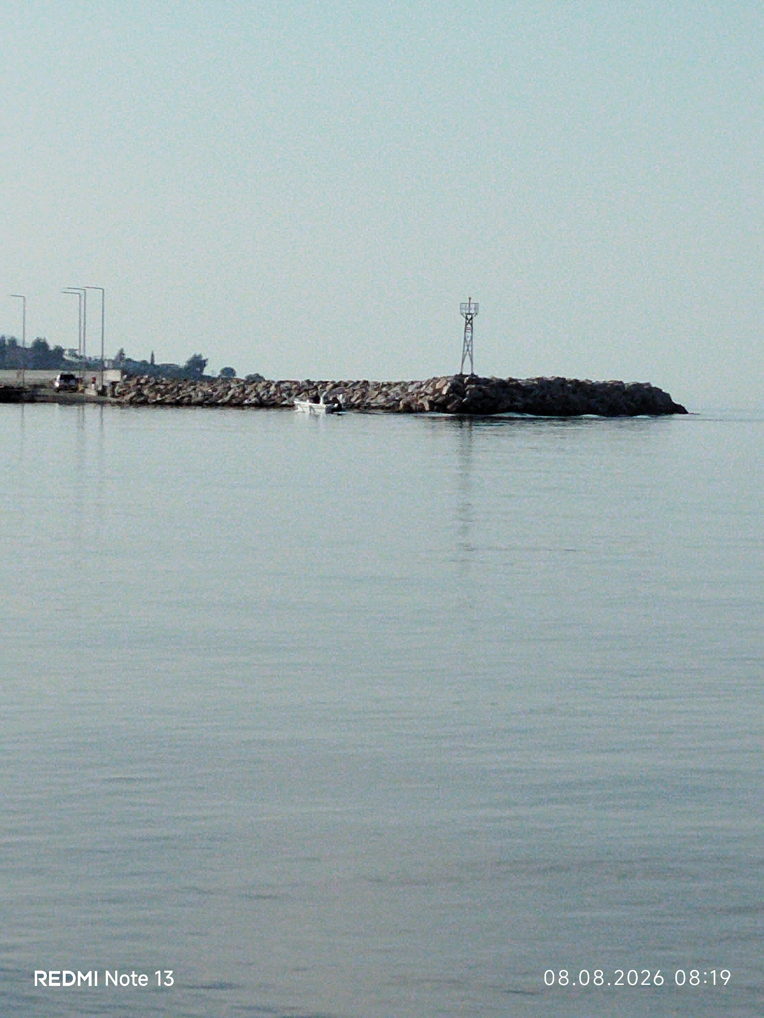 The image captures a serene coastal scene with a breakwater constructed of large rocks, extending into the calm, light blue sea. Atop the breakwater, a navigational aid with a distinctive cross-shaped top is visible. A small white boat with two people on board is navigating the water near the breakwater. In the distance, the shoreline of Sithonia Municipal Unit, Greece, is discernible, featuring trees and what appear to be utility poles or streetlights along a road. The overall atmosphere is tranquil, with the sky a pale, hazy blue, and the water reflecting the muted tones of the sky and surroundings. The lower portion of the image displays text overlays: "REDMI Note 13" on the left and "08.08.2026 08:19" on the right, indicating the device used for capture and the date and time.