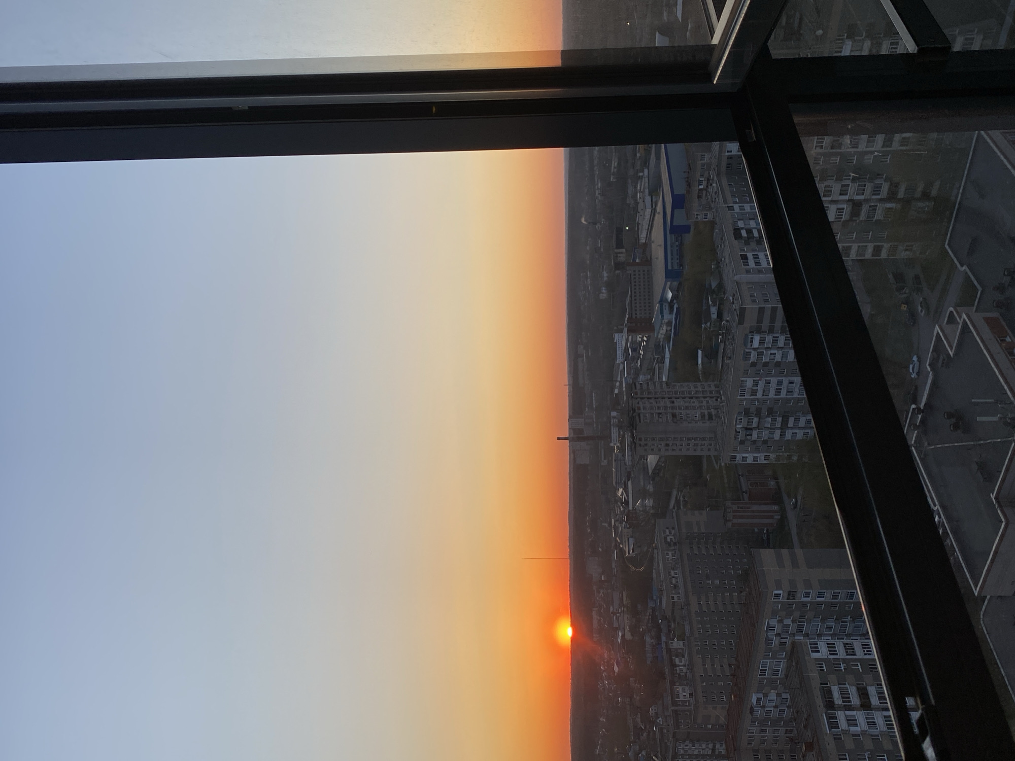 The image shows a sunset viewed from a high vantage point in Izhevsk, Russia. The sun is positioned low on the horizon, radiating an orange glow that is reflected in the sky and illuminates the urban landscape. The skyline consists of a mix of mid-rise buildings, including residential apartment blocks with multiple rows of windows. A tall tower is visible in the distance. The foreground is framed by the dark structure of a window or viewing platform. The image is taken from inside a building.