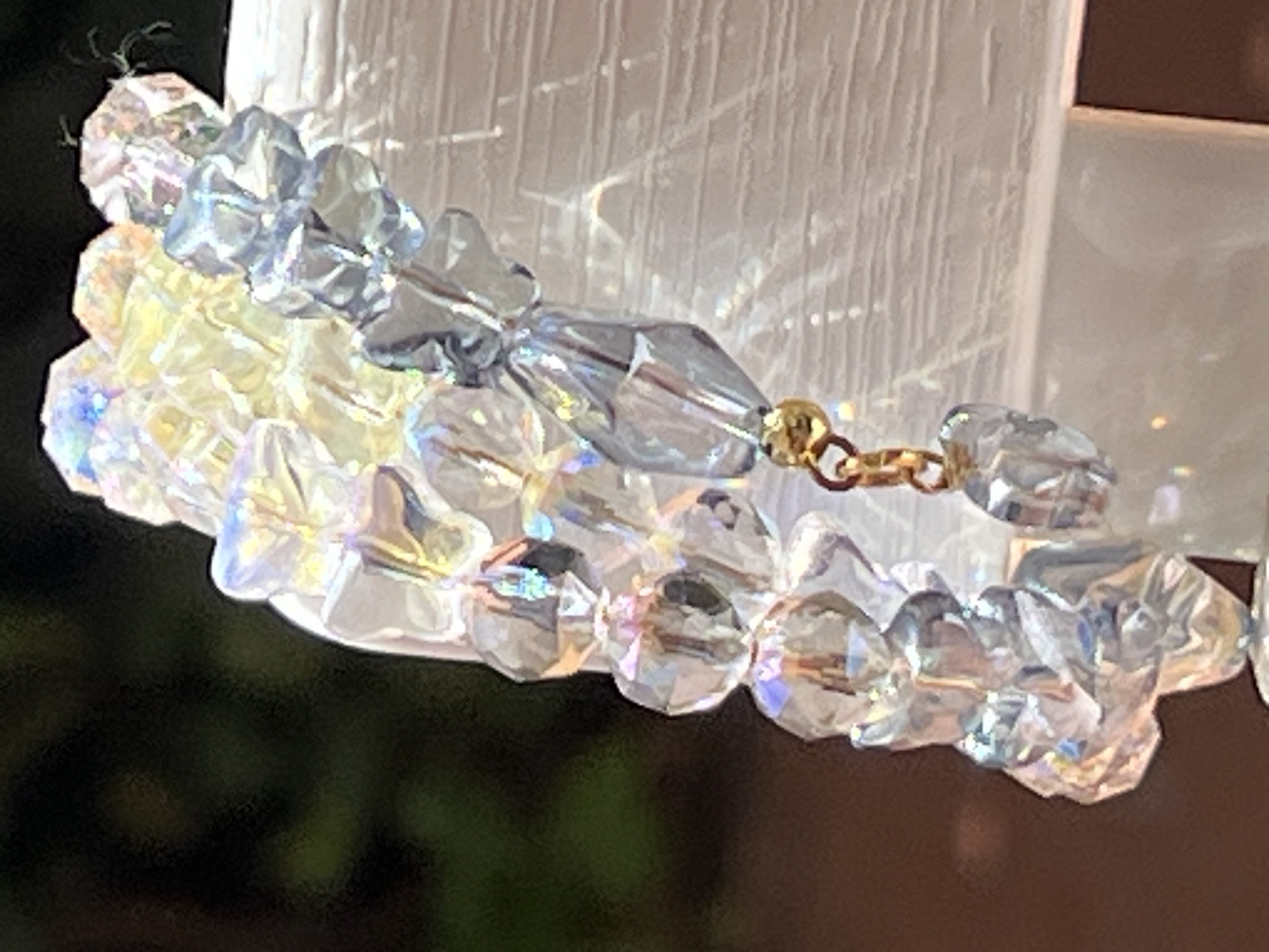 The image shows a close-up of a bracelet made of iridescent crystals. The bracelet is composed of various faceted beads, some appearing to be star-shaped and others more elongated. There is a small gold-colored charm or link attached to the bracelet. The crystals reflect light, creating a shimmering effect with hints of blue, yellow, and purple. The bracelet is resting on a white, textured surface, possibly a piece of furniture or a display stand. In the background, a dark, blurred area suggests foliage, indicating an outdoor setting, possibly a garden or a balcony. There is no identifiable city or location information present in the image itself, and therefore no relation to Bijela, Montenegro can be established.