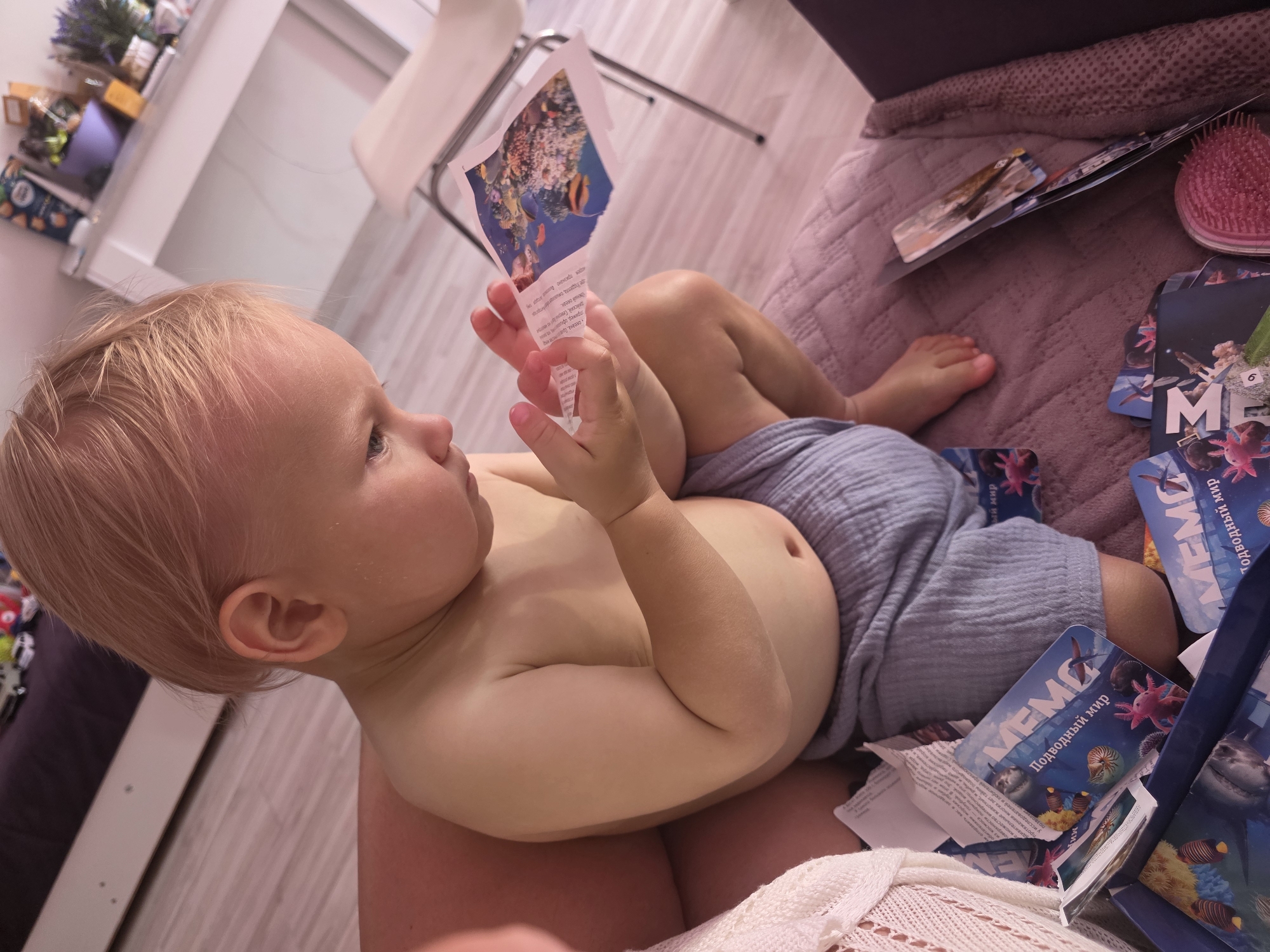 A young child with blonde hair lies on a textured purple blanket, looking at a torn piece of paper with an image of coral and fish. The child is wearing a light blue diaper or shorts. Around the child are several cards with the word "MEMO" and images of underwater creatures, suggesting a game or educational activity related to the ocean. One card shows an axolotl and the text "Подводный мир", which translates to "Underwater world". Another card has a shark and coral. A pink hairbrush is also visible on the blanket. In the background, part of a white cabinet with various items on it, including what looks like dried flowers, and a white chair are visible. The lighting suggests it is daytime. The presence of a hairbrush and children's items might indicate this is a home environment.