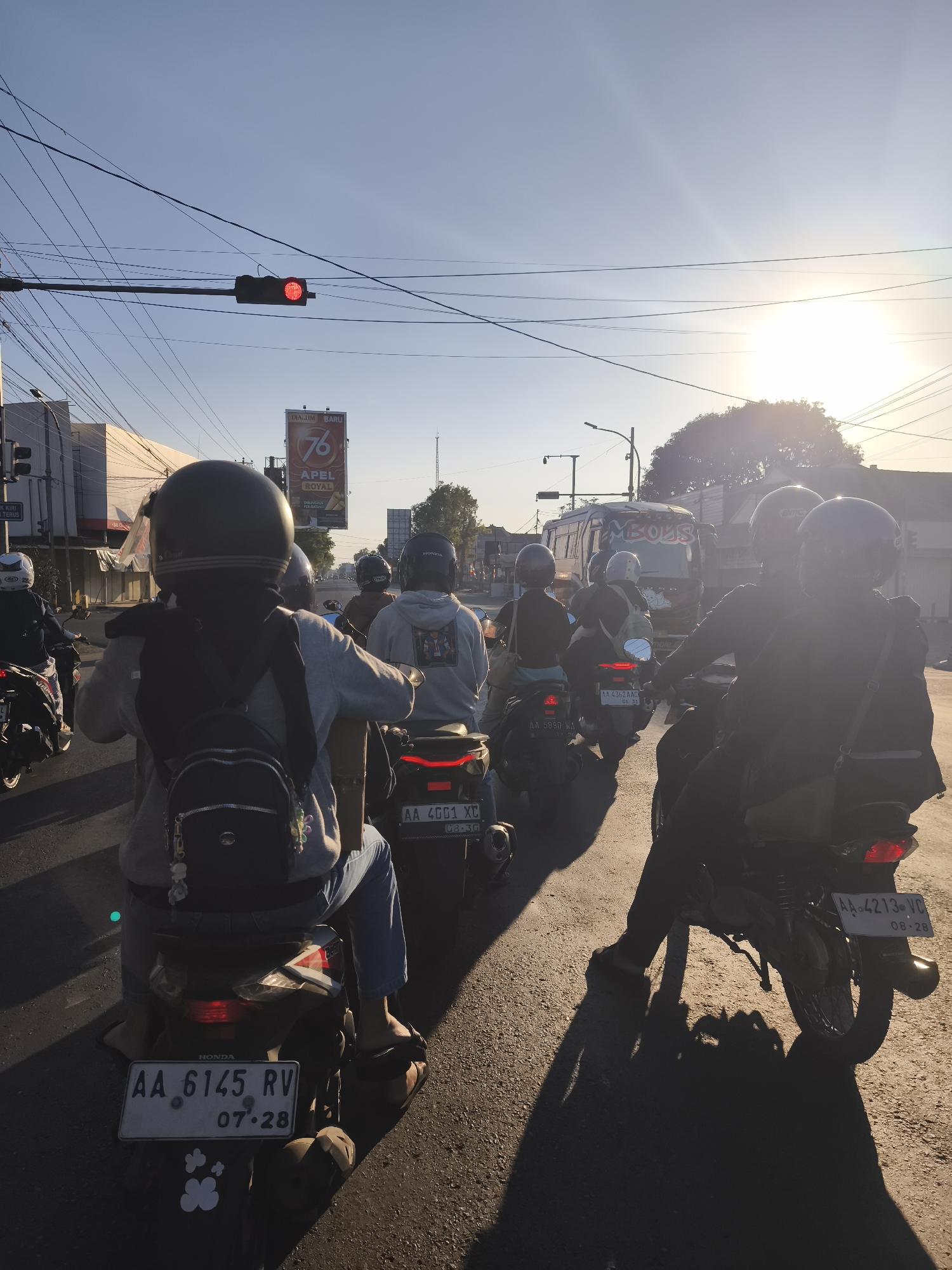 The image depicts a street scene in Kutoarjo, Indonesia, at dusk. A group of motorcyclists are stopped at a red traffic light. The sky is clear with the sun setting, casting long shadows across the asphalt road. The motorcyclists are wearing helmets and casual clothing. One motorcyclist in the foreground is wearing a grey hoodie and blue jeans, with a black backpack. Another motorcyclist to the right is wearing a dark hoodie. In the background, there is a bus with "AMBOLS" written on its side and a billboard for "76 APEL ROYAL". The traffic light is red, indicating that vehicles must stop. The road is paved and appears to be in good condition. The overall scene suggests a typical urban commute during the evening hours.