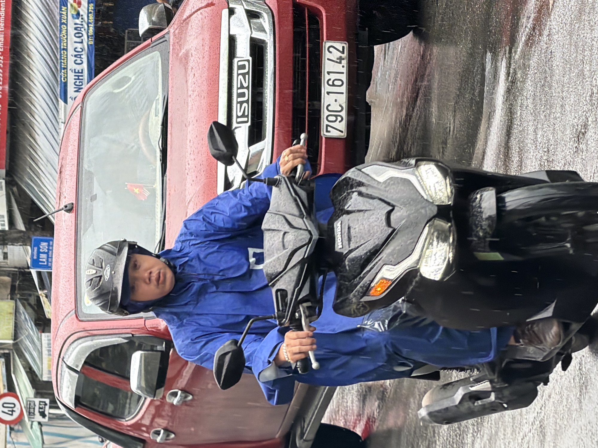 A person wearing a blue rain suit and a black helmet rides a black scooter on a wet street in Nha Trang, Vietnam. Rain is falling, creating reflections on the road. A red Isuzu pickup truck is visible behind the scooter, with its license plate reading "79C-161.42". Various signs are present in the background, including a speed limit sign displaying "40" and a blue sign with Vietnamese text. The overall scene depicts urban traffic during a rainy day.