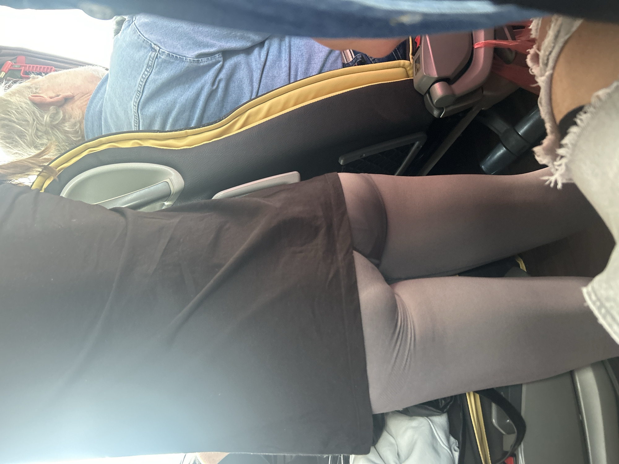 The image captures an interior scene, likely inside a bus or similar public transport vehicle, with a vantage point from behind a seat looking forward. In the foreground, partially obscured, is the upper back of a dark-colored seat. The primary focus is on the lower body of a person seated closer to the camera, wearing a black top or dress over light gray opaque tights or leggings. Their legs and the curve of their hip are visible. To the far right, part of another person's leg and foot, clad in ripped light-colored fabric (possibly shorts or jeans), can be seen.
Further back, beyond the seat in the foreground, an older individual with light gray, curly hair and a visible ear is seen from the side, appearing to be asleep or resting. They are wearing a light blue denim shirt. This person is occupying a dark-colored seat with a distinctive yellow fabric trim along its top edge. An armrest with a silver-colored handle or cup holder is prominent on this seat. Below it, a small white rectangular object rests on the armrest. Red and black straps or belts are visible near the seat mechanisms. The overall lighting suggests it is daytime. No text is visible within the frame. The scene suggests travel, potentially in or around Gravouna, Greece.