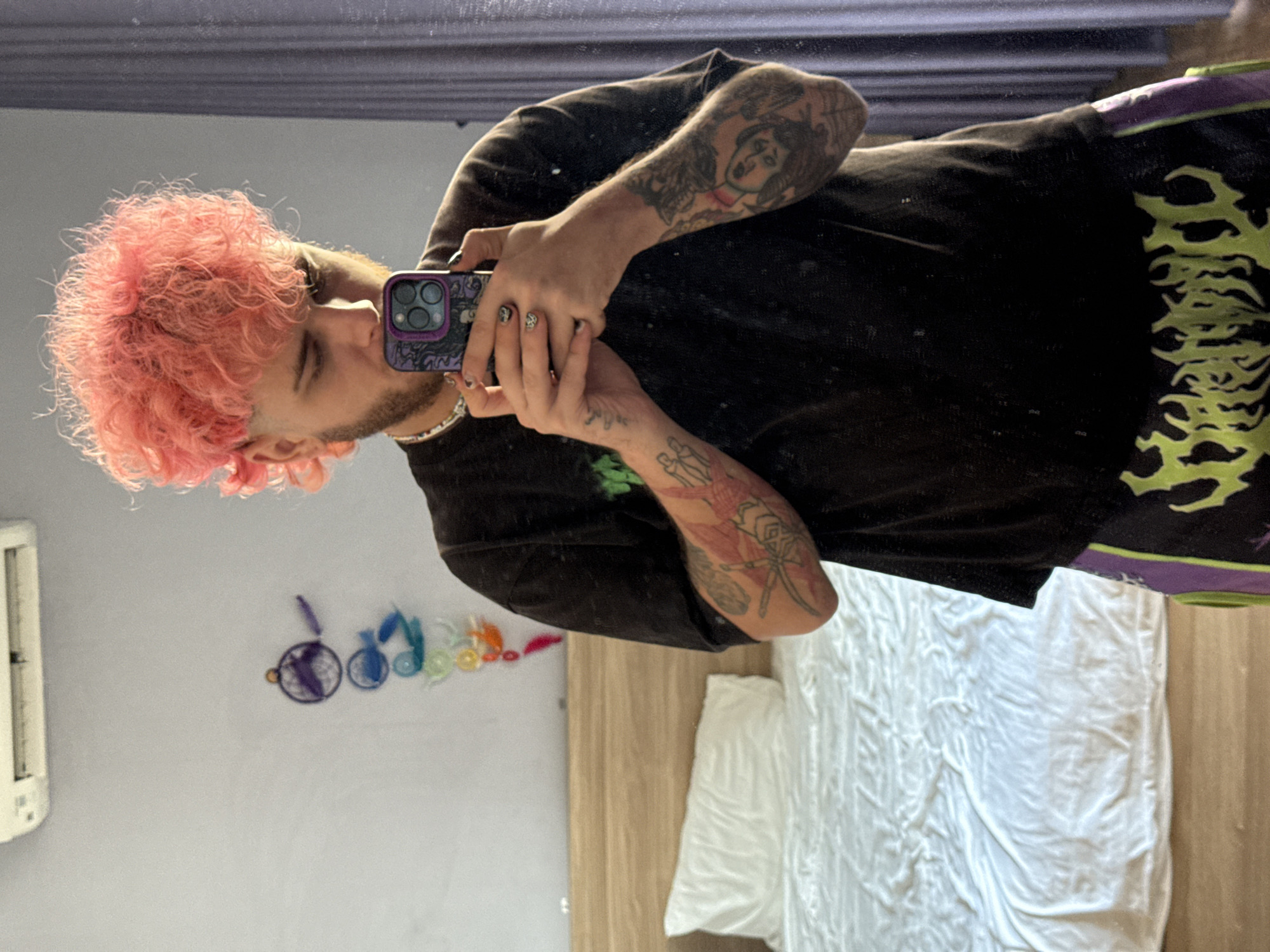 A person with pink hair and multiple tattoos is taking a selfie in a mirror. The person is wearing a black shirt and shorts with a green and black pattern. The background includes a white bed, a wooden floor, and a wall with colorful decorations. An air conditioning unit is visible on the wall. There is no indication of this image being taken in Đà Nẵng, Vietnam.