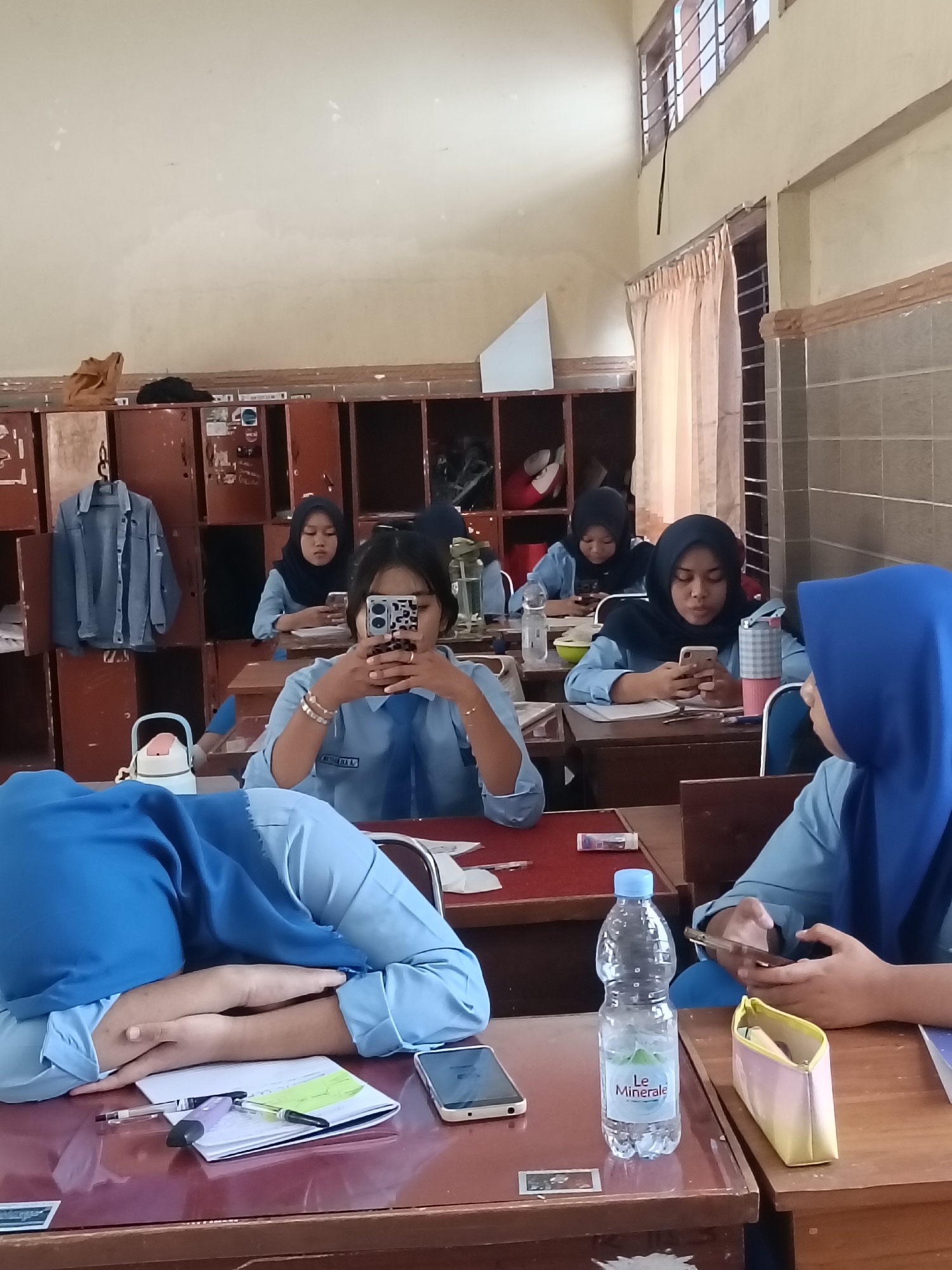 The image depicts a classroom scene in Kediri City, Indonesia, likely during school hours. Several young women, appearing to be students, are seated at desks. Most are wearing light blue uniforms with darker blue headscarves. Some are engaged with their mobile phones, while one student in the foreground is asleep with her head resting on her arms on the desk. Visible objects include water bottles, notebooks, pens, a pencil case, and a denim jacket hanging on a locker. The classroom environment appears somewhat dated, with wooden lockers and simple desks. The lighting suggests it is daytime, possibly with natural light entering from windows not fully visible.