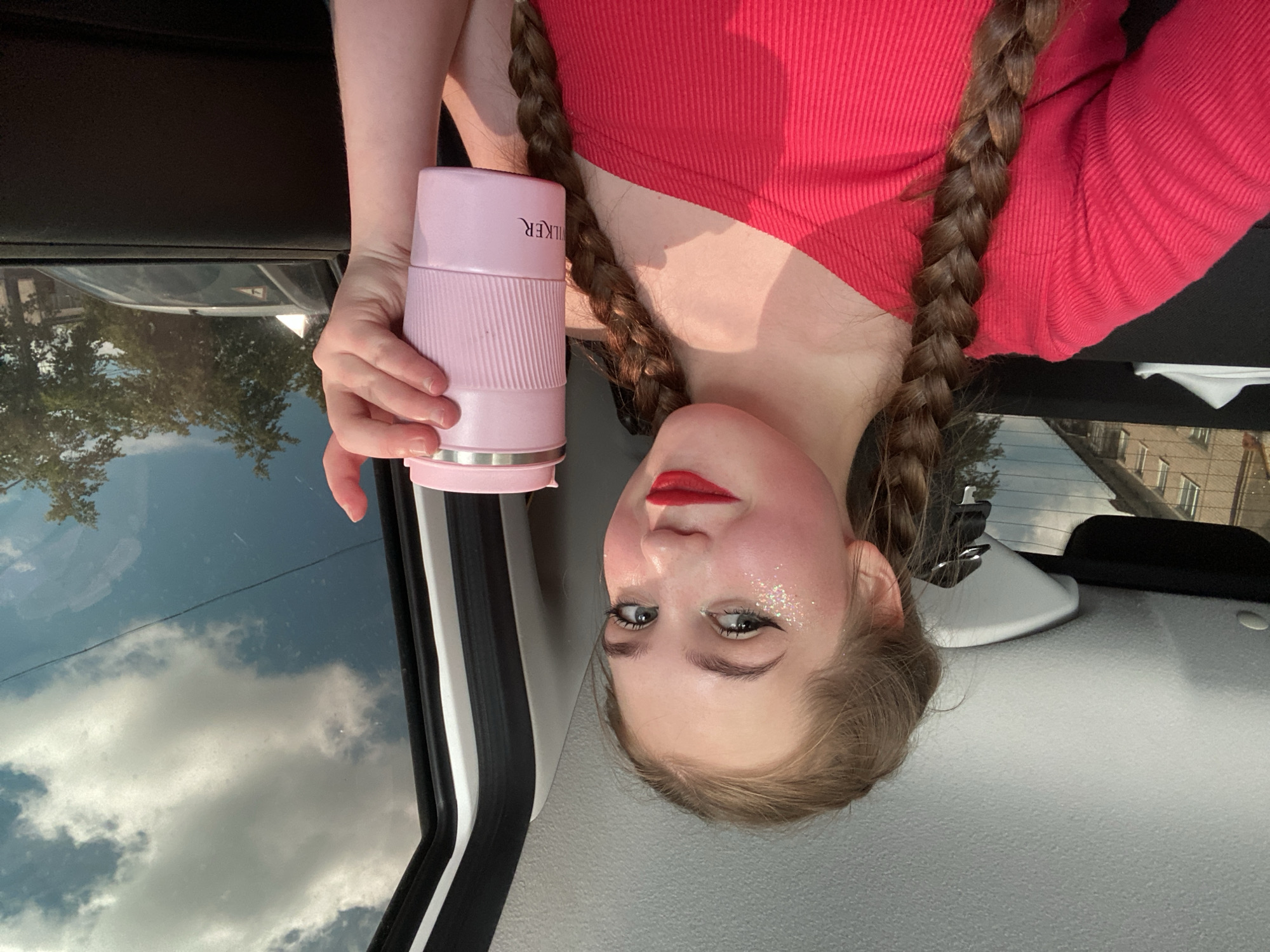 A young woman with blonde hair in two braids, wearing a red one-shoulder top, is holding a pink travel mug. She has makeup on, including red lipstick and glitter on her cheeks and eyelids. She is inside a vehicle, possibly a car, as indicated by the interior features and the view out the window. Outside the window, there are trees, buildings, and a cloudy sky, suggesting an urban or suburban environment. The image appears to be a selfie taken by the woman. The presence of the buildings visible through the window suggests a location in the city of Chaikovsky, Russia, given the context provided.
