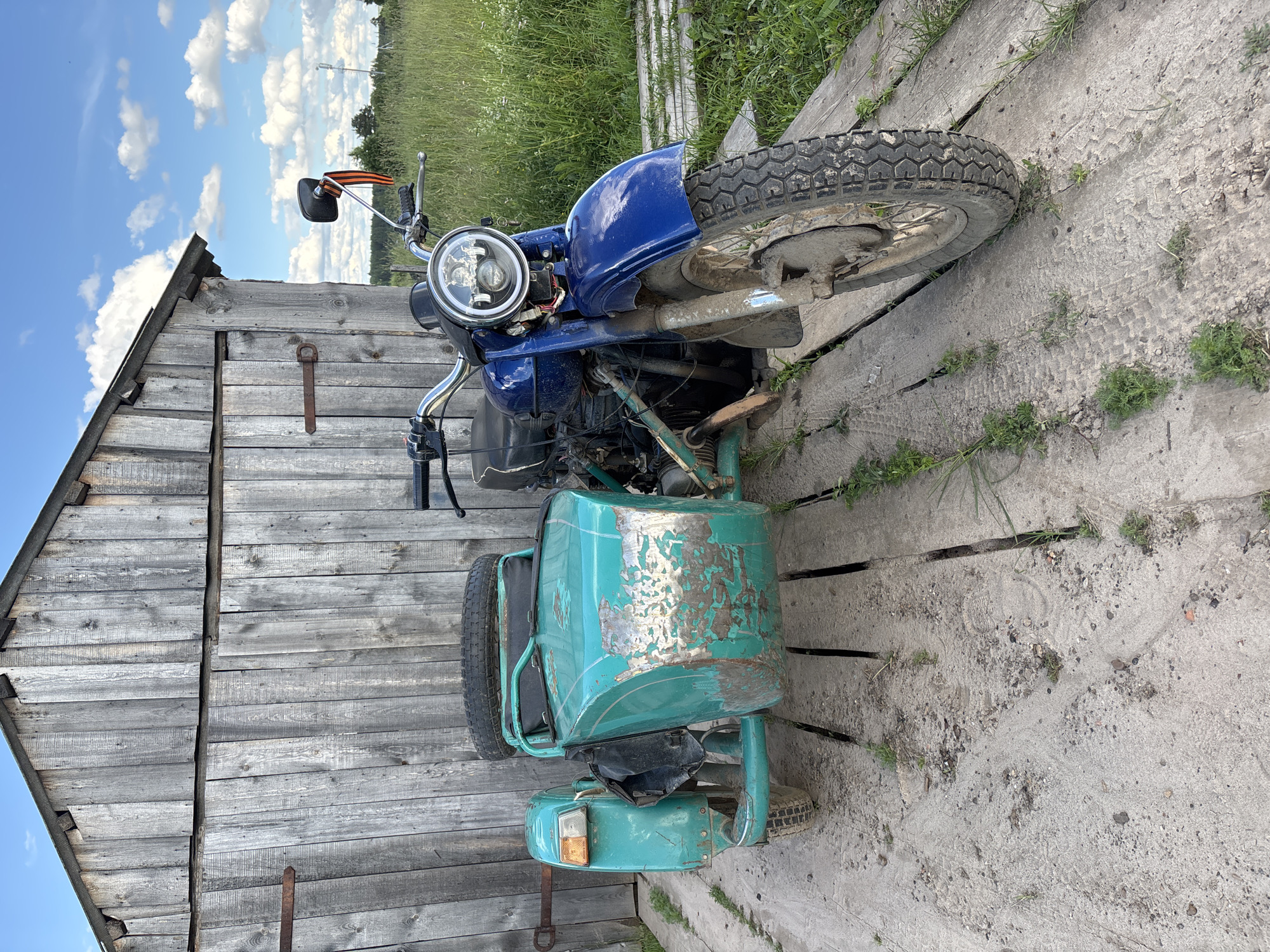 This media file showcases a blue and teal motorcycle with a sidecar, parked outdoors. The motorcycle appears to be older, with some wear and tear, particularly on the sidecar's paint. It is positioned on a wooden platform that looks like a ramp or walkway, next to a weathered wooden shed. The scene is set in a rural or semi-rural environment, with tall green grass and trees visible in the background under a bright blue sky dotted with white clouds. The lighting suggests it's daytime, likely late morning or afternoon. The overall impression is one of a rustic, possibly forgotten setting. There are no people visible in the image. The sidecar has a distressed teal finish with patches of silver showing through, indicating rust or missing paint. The motorcycle has a round headlight and chrome handlebars. A striped ribbon, possibly of military significance, is attached to one of the handlebars. The wooden shed is constructed from rough, weathered planks. The ground is a mix of dirt and what appears to be a concrete or wooden surface with some cracks and vegetation growing through. The location context of Kurgan, Russia, suggests a setting within that region.