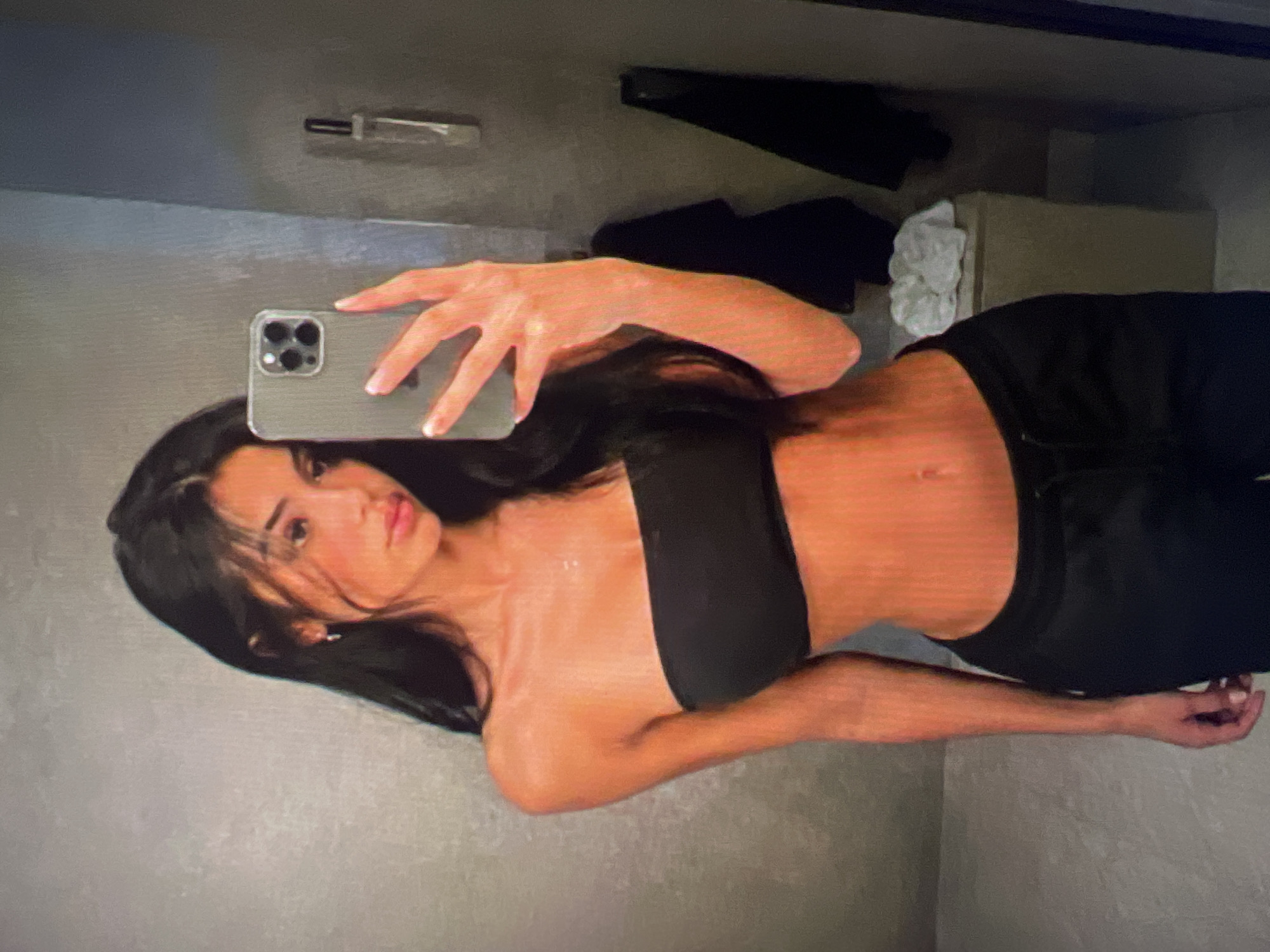 A young woman with long, dark hair takes a mirror selfie in what appears to be a bathroom or dressing room. She is wearing a black strapless top and black sweatpants. Her phone, a silver iPhone with a protective case, is held up to take the photo. The background shows a textured gray wall, a small shelf with a bottle, and dark clothing hanging on a rack. There is also a light-colored object, possibly a towel or clothing, on a counter to the right.