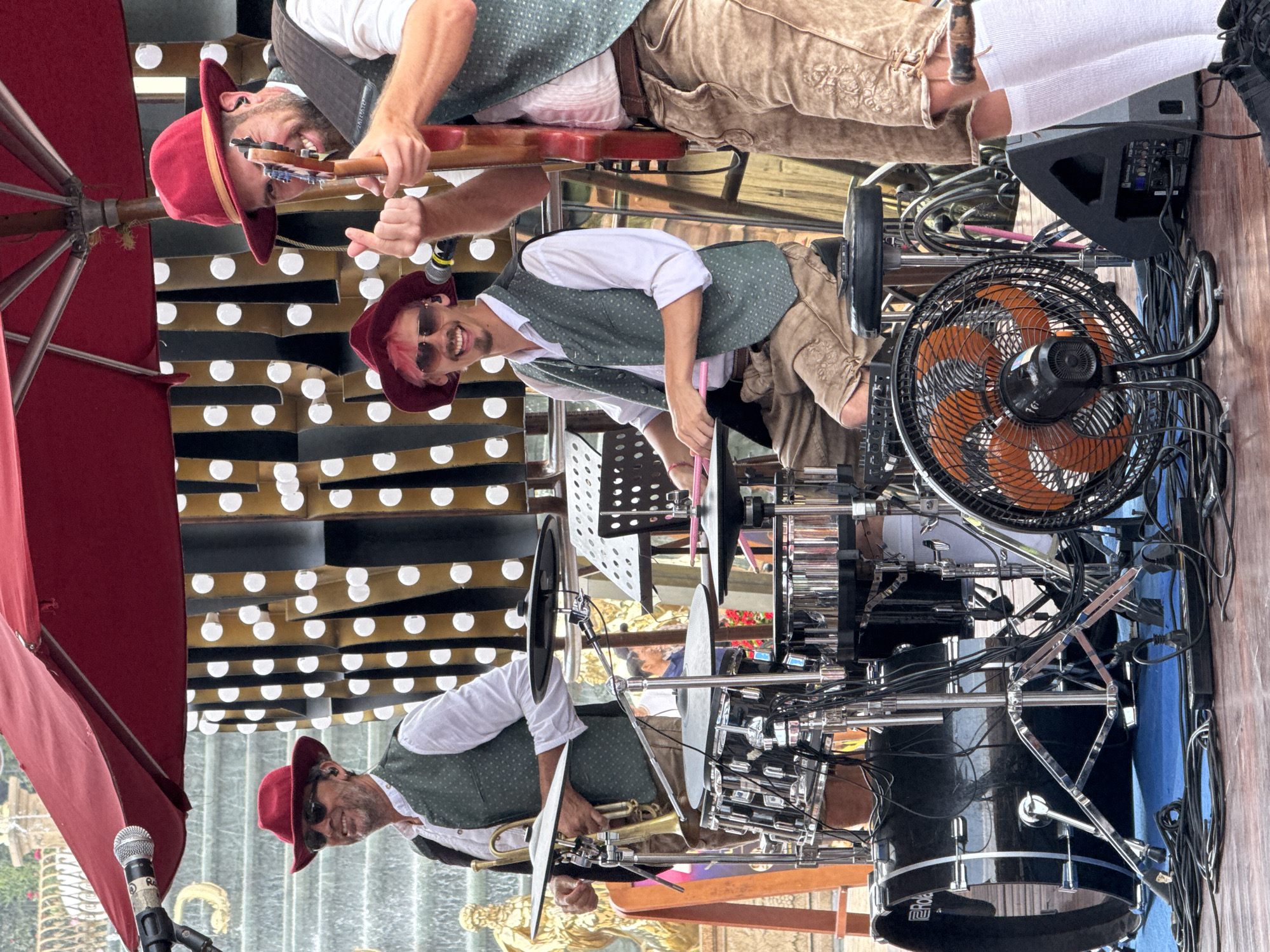 The image shows three musicians in traditional Bavarian outfits playing instruments outdoors in what appears to be Đà Nẵng, Vietnam. The musicians are wearing red hats, white shirts, green vests, and tan lederhosen. One musician is playing a guitar, another is playing a drum set, and the third is playing a trumpet. An orange fan is next to the drum set. A red umbrella is to the left, and a waterfall background featuring decorative lights is visible behind them.