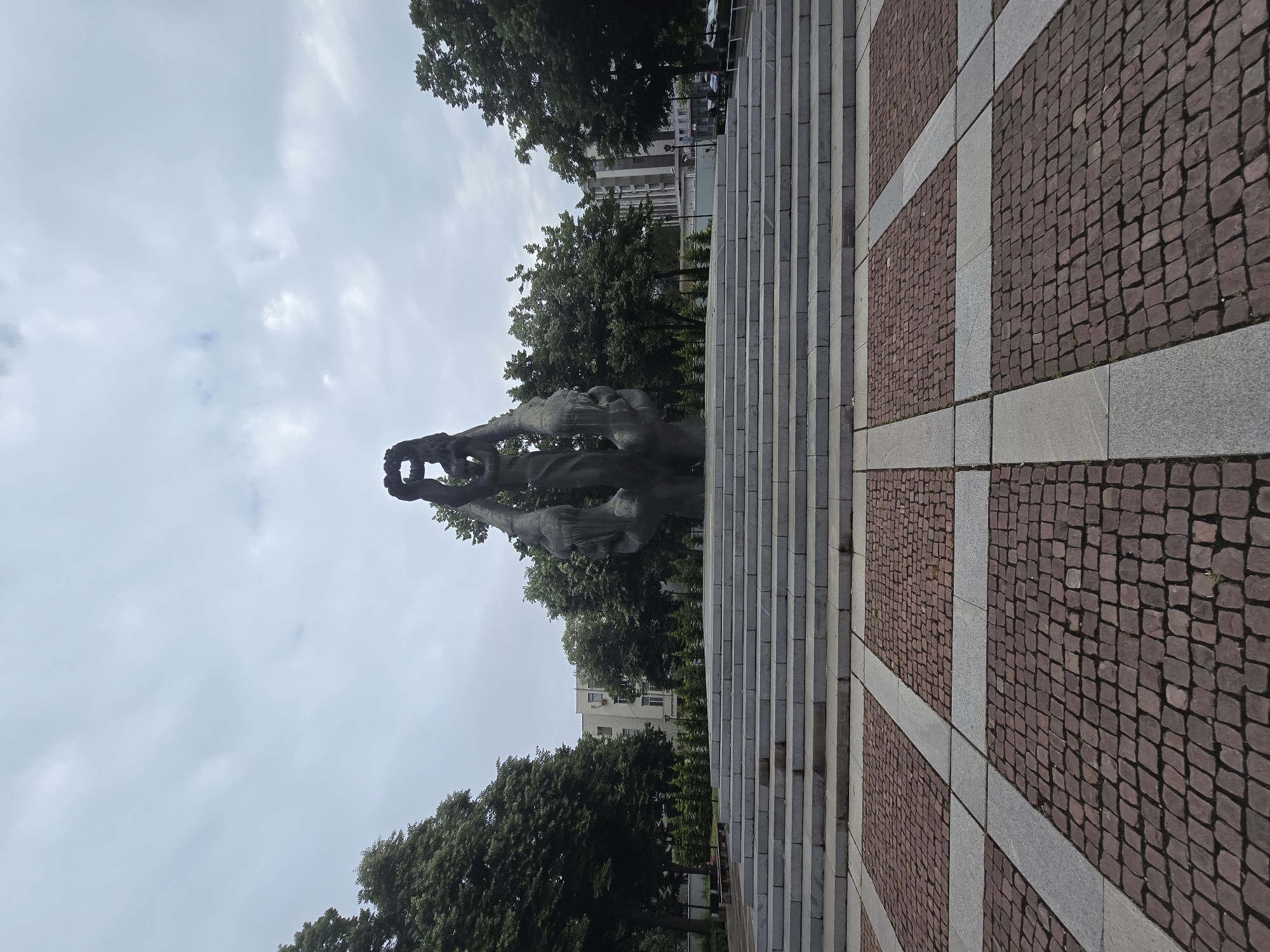The media file displays an outdoor scene, likely a public square or park, under an overcast sky. The central subject is a large, dark, monumental statue featuring multiple figures. These figures appear dynamic, possibly reaching upwards or holding an object together, with one figure prominently raising something overhead.
To the right of the statue, a wide, grey stone staircase ascends, leading towards unseen features at the top. Adjacent to the staircase, on the right side of the frame, is a paved area with a distinct pattern. This paving consists of square sections of reddish-brown cobblestones, separated and outlined by lighter grey stone strips, creating a grid-like design on the ground.
In the background, behind the statue and staircase, numerous lush green trees are visible, suggesting a park-like environment. Beyond the trees, glimpses of multi-story buildings can be seen. The sky is largely covered by light grey clouds, indicating an overcast day. No specific activity or event is taking place in the scene, which appears static. The time of day is daytime, but without direct sunlight. There is no visible text in the image.