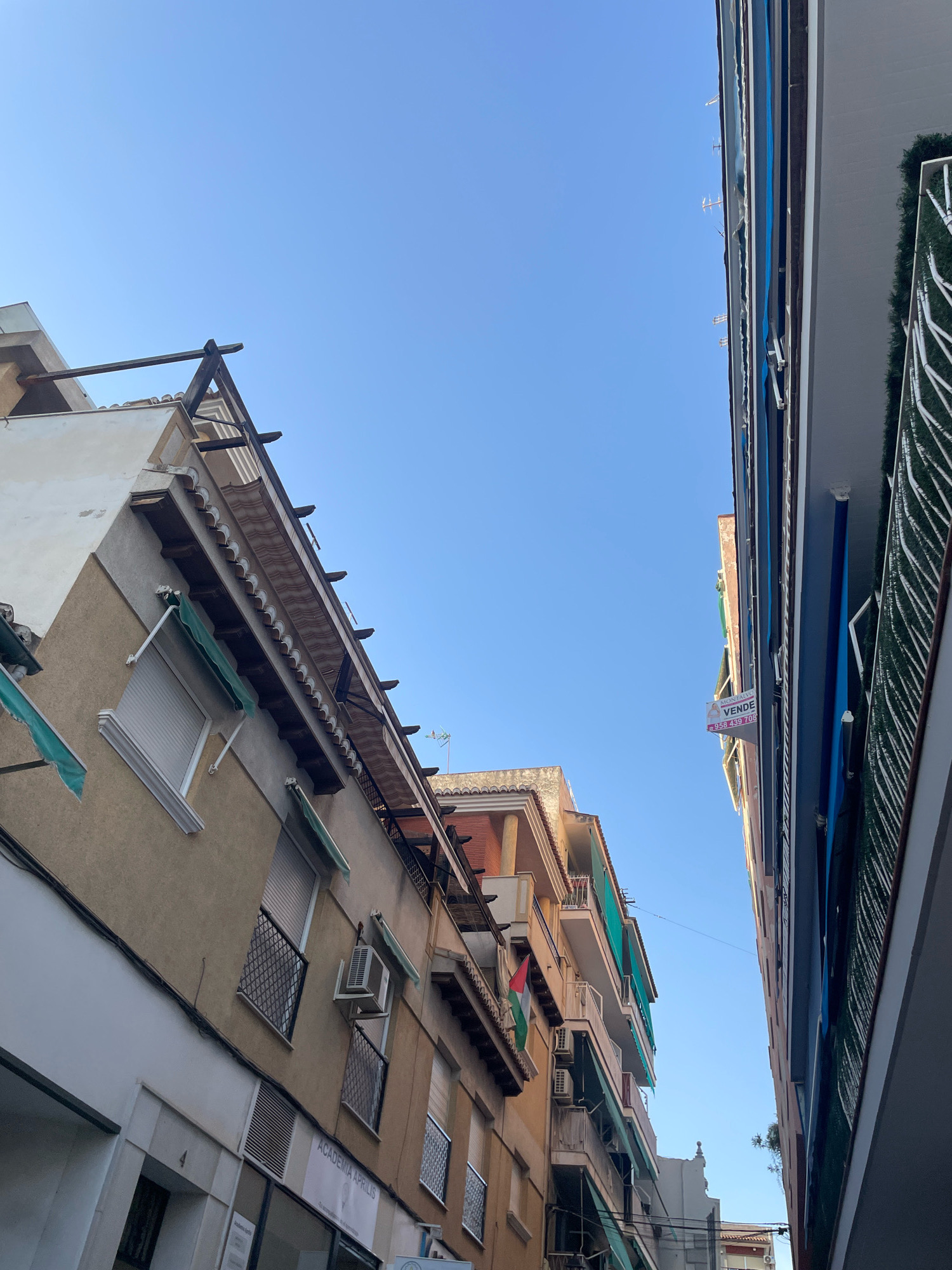 The image shows a narrow street in Atarfe, Spain, with buildings on both sides. The sky is clear and blue. On the left side of the street, there is a building with a beige facade, featuring several windows with green awnings and white rolling shutters. An air conditioning unit is visible on the facade. The building also has a balcony with a decorative railing and a flag hanging from it. There is a sign for "ACADEMIA APRILIS" above a ground-floor storefront.
On the right side of the street, another building is visible, with a blue facade and a balcony with green fencing. A "VENDE" (For Sale) sign with a phone number is attached to this building. The buildings appear to be old, with architectural details such as decorative cornices and tiled roofs. The perspective is looking upwards, emphasizing the height of the buildings and the narrowness of the street. The lighting suggests it is daytime, with shadows cast by the buildings.