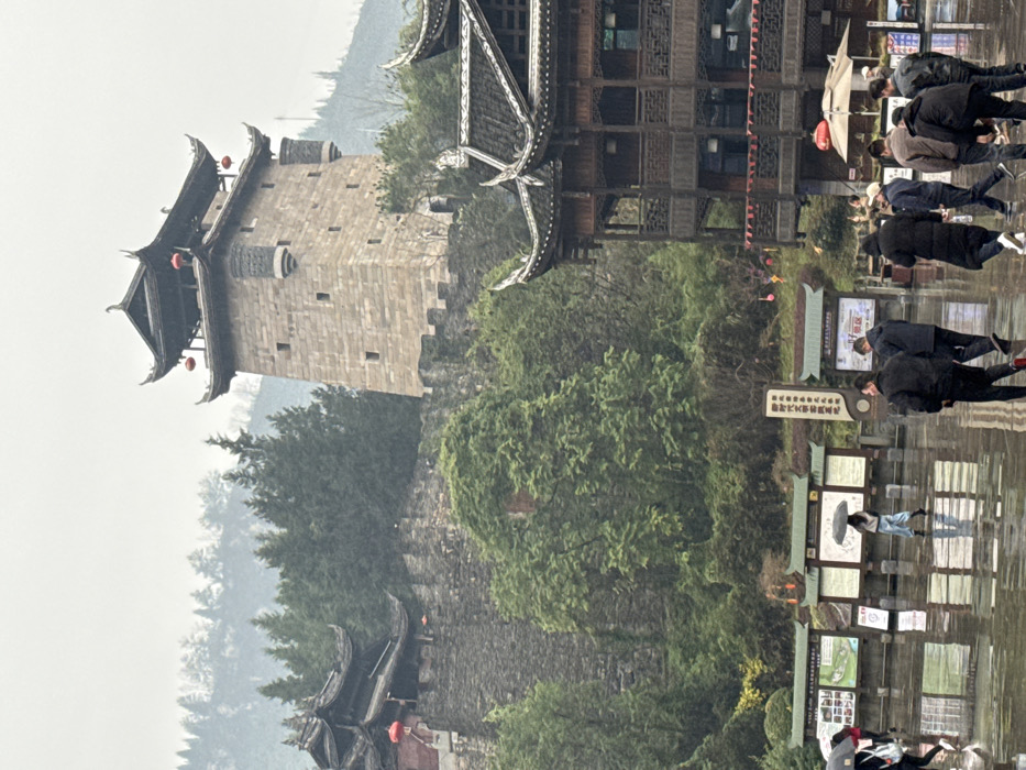 The image depicts a traditional Chinese architectural complex situated in Pengshui Miao and Tujia Autonomous County, China, under an overcast sky with evidence of rain.
In the upper-middle and left sections, a series of ancient-style structures ascend a vegetated hillside. A prominent, four-story stone watchtower, featuring light-colored masonry, crenelated battlements, and two projecting side turrets, dominates the upper-right portion. It is capped with a dark, multi-tiered traditional Chinese roof adorned with red lanterns hanging from its eaves. To the left of this tower, a substantial dark stone wall with foliage extends horizontally, supporting another traditional dark-roofed building, partially visible, also featuring red lanterns. The hillside itself is densely covered with green trees and undergrowth, particularly prominent behind and around the stone walls.
In the foreground, a wide, dark paved area, reflecting the sky and surrounding objects due to a wet surface, occupies the lower half of the frame. Scattered across this wet ground are approximately 10-15 individuals. One person in the mid-ground, dressed in light attire, walks alone holding a black umbrella. Several other figures, primarily men dressed in dark clothing, walk in small groups or individually in the lower-right foreground. To the left and center of the mid-ground, a series of large information boards with green-tiled roofs and white display panels, containing Chinese text and graphics, are arranged in a row. A partial view of a multi-story traditional wooden building with intricate latticework and a similar dark, tiered roof structure is visible in the lower-right, bordering the wet plaza. The overall atmosphere is muted, consistent with cloudy and wet weather conditions.