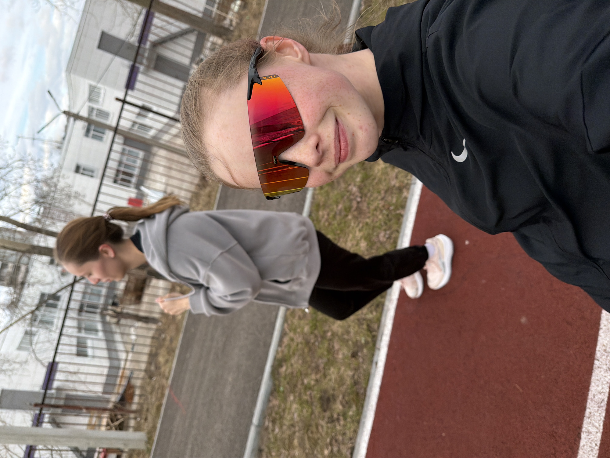 Two young women are captured in a selfie-style photograph on an outdoor athletic track, likely in Berezniki, Russia, during what appears to be a cool, overcast day in late winter or early spring.
The subject closest to the camera is taking the selfie. She has light hair, visible freckles across her nose and cheeks, and a slight smile. She is wearing large, sporty sunglasses with highly reflective orange and red lenses, on which the brand "SJO" is visible on the frame. Her attire includes a black athletic jacket with a white Nike swoosh logo on the left chest.
Further in the background, another young woman is visible, in motion or preparing for activity. She has darker hair pulled back into a ponytail and is wearing a light grey hooded sweatshirt and dark pants. Her feet, clad in light-colored sneakers, are on the red track. She appears to be holding a white cord or band in her hands.
The setting includes a reddish-brown athletic track with white lane markings, bordered by a strip of dry, patchy grass. In the mid-ground, a metal fence separates the track from a multi-story white building with numerous windows, suggesting a school or sports complex. Several bare trees are scattered in the background, reinforcing the seasonal inference. The sky is overcast and bright.