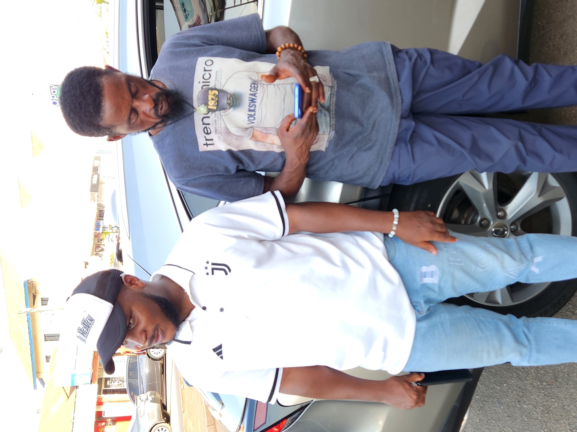 Two men are standing in front of a silver car. One man is wearing a gray shirt with a graphic design, blue pants, and a wooden beaded bracelet, and he is looking at a blue smartphone. The other man is wearing a white polo shirt with black trim, light blue jeans with white lettering, and a black and white baseball cap. The setting appears to be an urban area, possibly a city, with buildings and parked cars visible in the background.