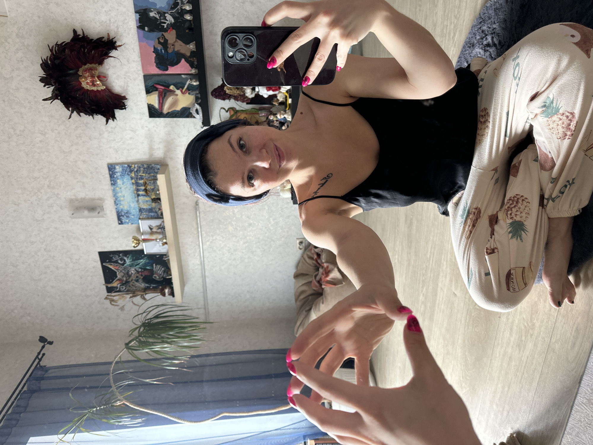 The image shows a woman taking a selfie in a room. She is wearing a black tank top, patterned pajama pants, and a dark blue headband. She is holding a black smartphone with a triple-lens camera. Her nails are painted bright pink.
Behind her, on the wall, are various decorative items, including a feathered headdress and several framed pictures or artwork. A shelf with various objects is visible.
To the left of the frame, there is a plant near a window with blue curtains. In the reflection, another person can be seen.
The floor is light-colored wood. Based on the available information, there's insufficient data to confirm the location in the city of Vladimir, Russia.