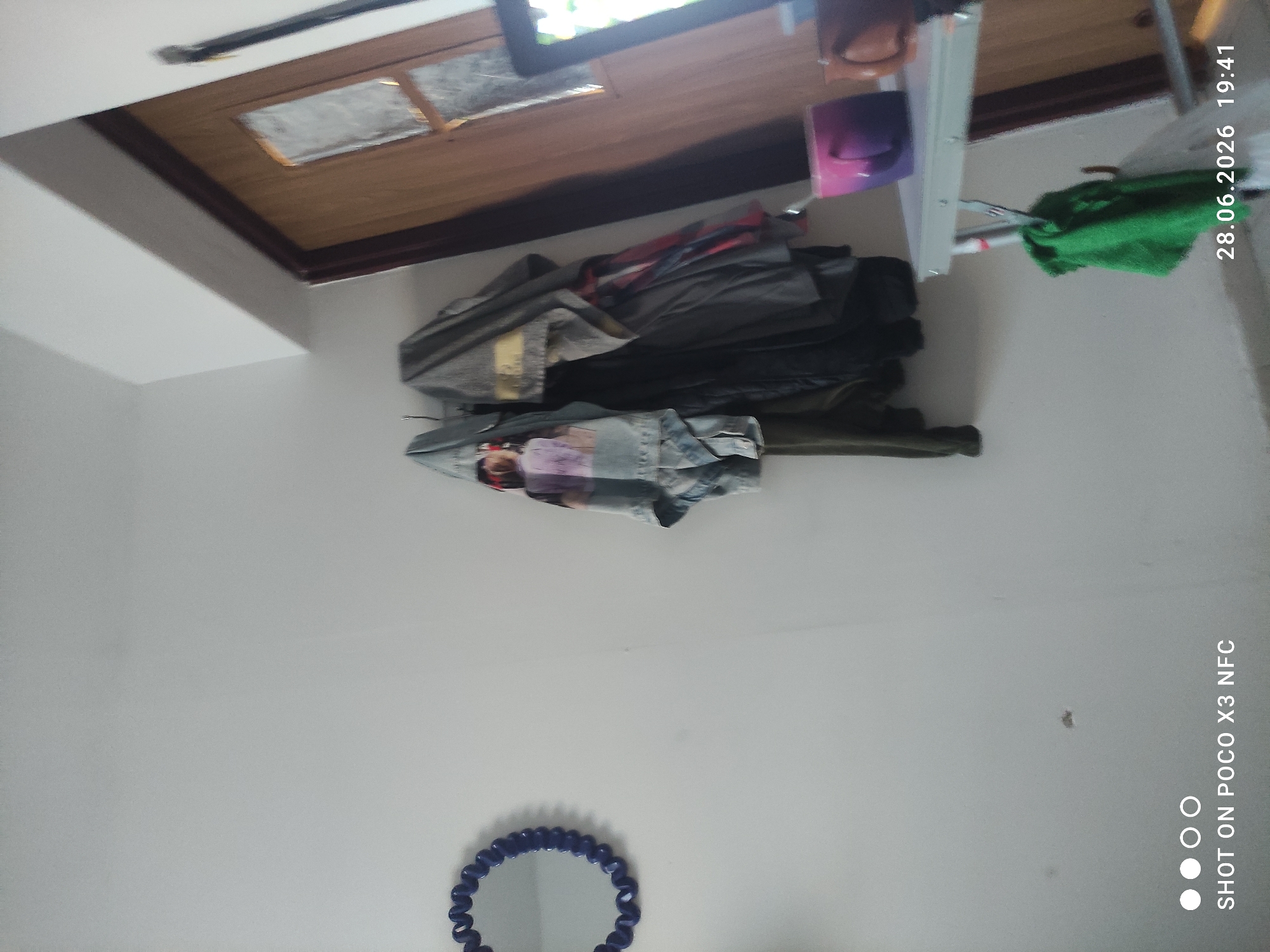In a room with white walls, a blue, circular mirror with a decorative edge hangs on the left. To the right of the mirror, several pieces of clothing are hung on a rack, including a denim jacket with a colorful graphic on the back, a gray jacket, and other garments. A doorway with a wooden door and a glass pane is visible on the right side of the image. In the foreground, a white metal table is set up, holding a purple mug and a brown mug. A green cloth is hanging from the table. The timestamp "28.06.2026 19:41" is displayed in the bottom right corner, along with the text "SHOT ON POCO X3 NFC". The scene appears to be indoors, likely in a residential setting.