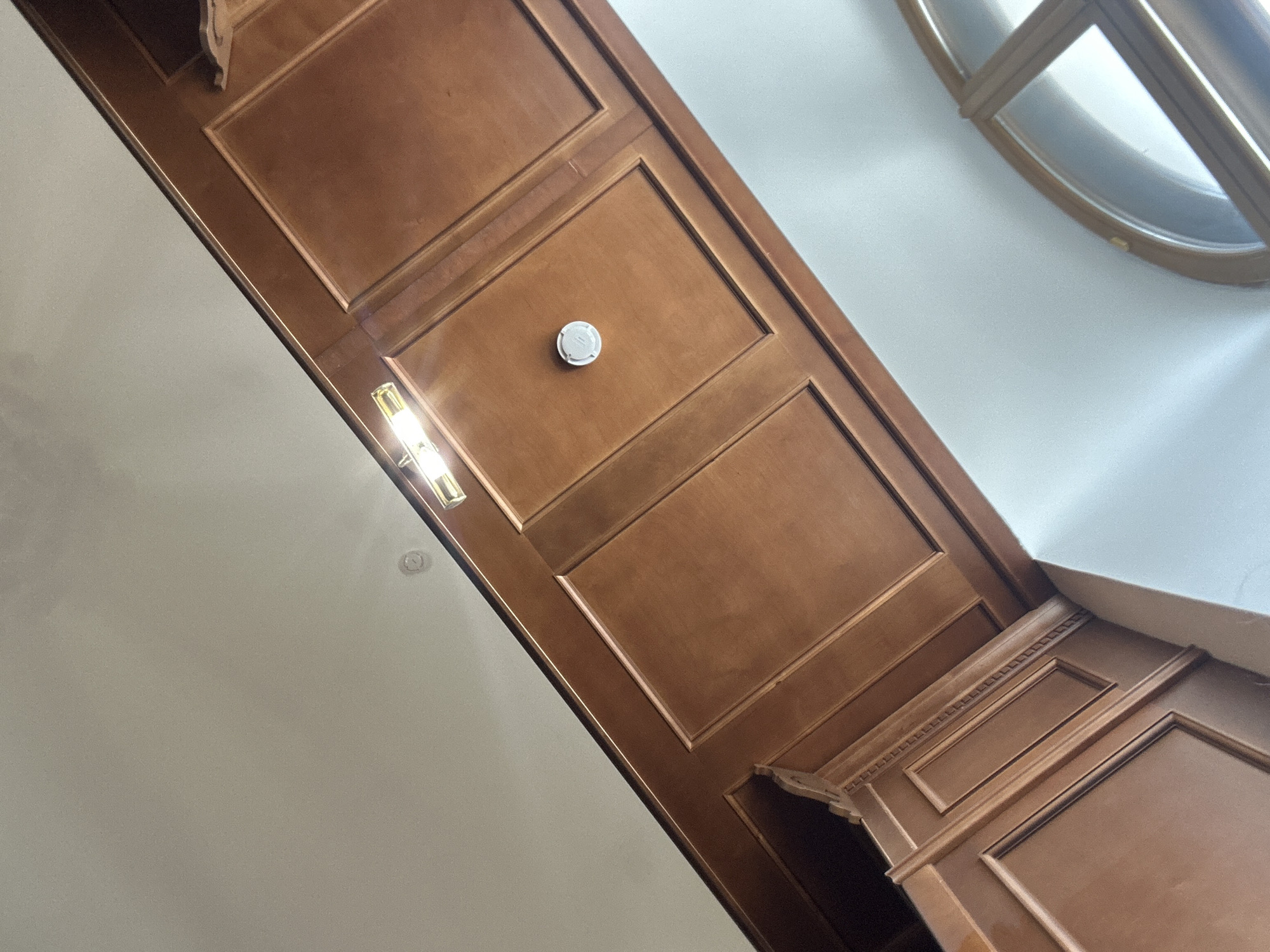 The image shows an interior view of a room with wood paneling on the ceiling and walls. The wood is a warm, medium-brown color with a polished finish. The ceiling has recessed panels, and a linear light fixture with two bulbs is visible. A round smoke detector is mounted in one of the ceiling panels. The wall also features wood paneling with decorative molding. In the background, a portion of an arched window with wooden mullions is visible. The lighting in the room is bright, casting a warm glow. The overall impression is one of traditional and elegant interior design.