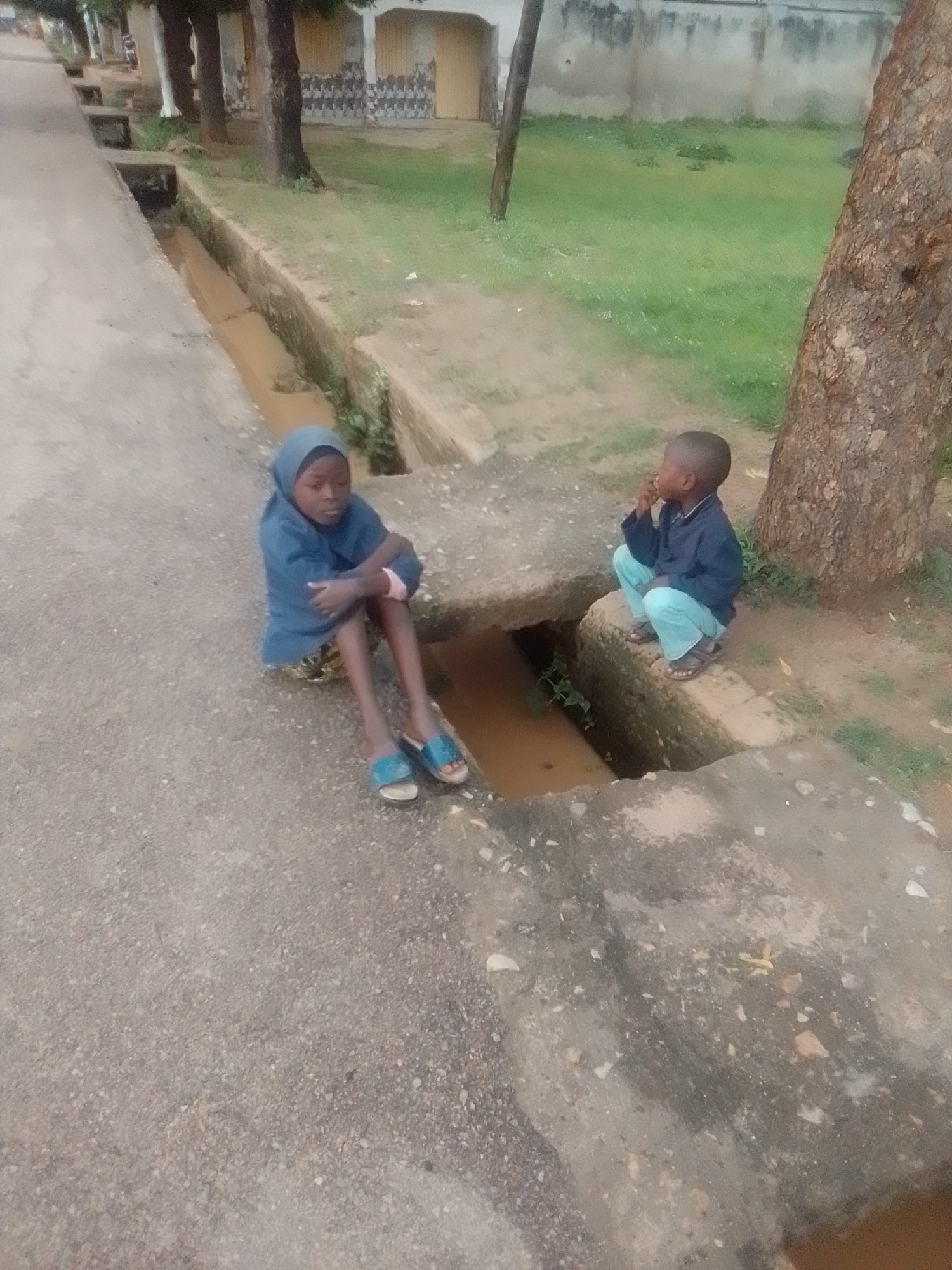 Two children, a girl and a boy, are present in this scene, likely in the city of Gombe, Nigeria. The girl, wearing a blue headscarf and a blue long-sleeved garment, sits on the edge of a concrete drainage ditch, her legs outstretched. She is wearing blue sandals. The boy, dressed in a dark blue long-sleeved shirt and light blue pants, is crouched on a concrete ledge adjacent to the ditch, with his right hand near his face. The ditch contains murky water and some vegetation. The surrounding environment consists of a paved surface, a grassy area with trees, and a building in the background. The lighting suggests it is daytime.